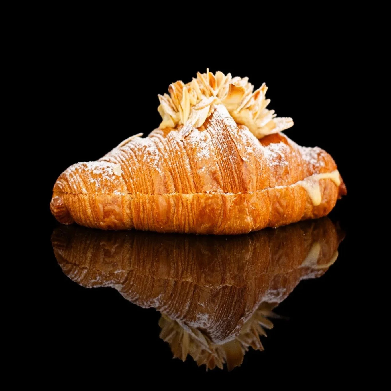 Un croissant con azúcar y almendras fileteadas en la parte superior, reflejado en una superficie negra. Contenido de redes sociales.
