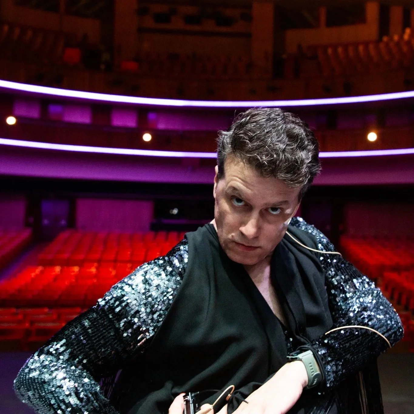 Hombre con expresión intensa en un teatro vacío con asientos rojos y luces púrpuras. Contenido de redes sociales.
