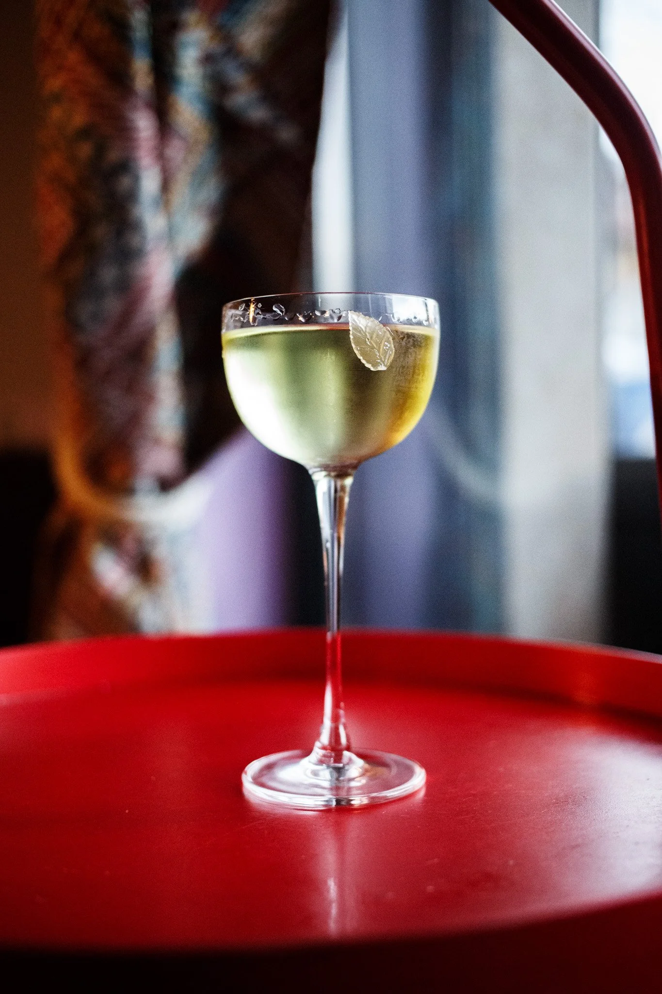 Copa de cóctel con una rodaja de limón en una mesa roja junto a una cortina y ventana en el fondo. Contenido de redes sociales.