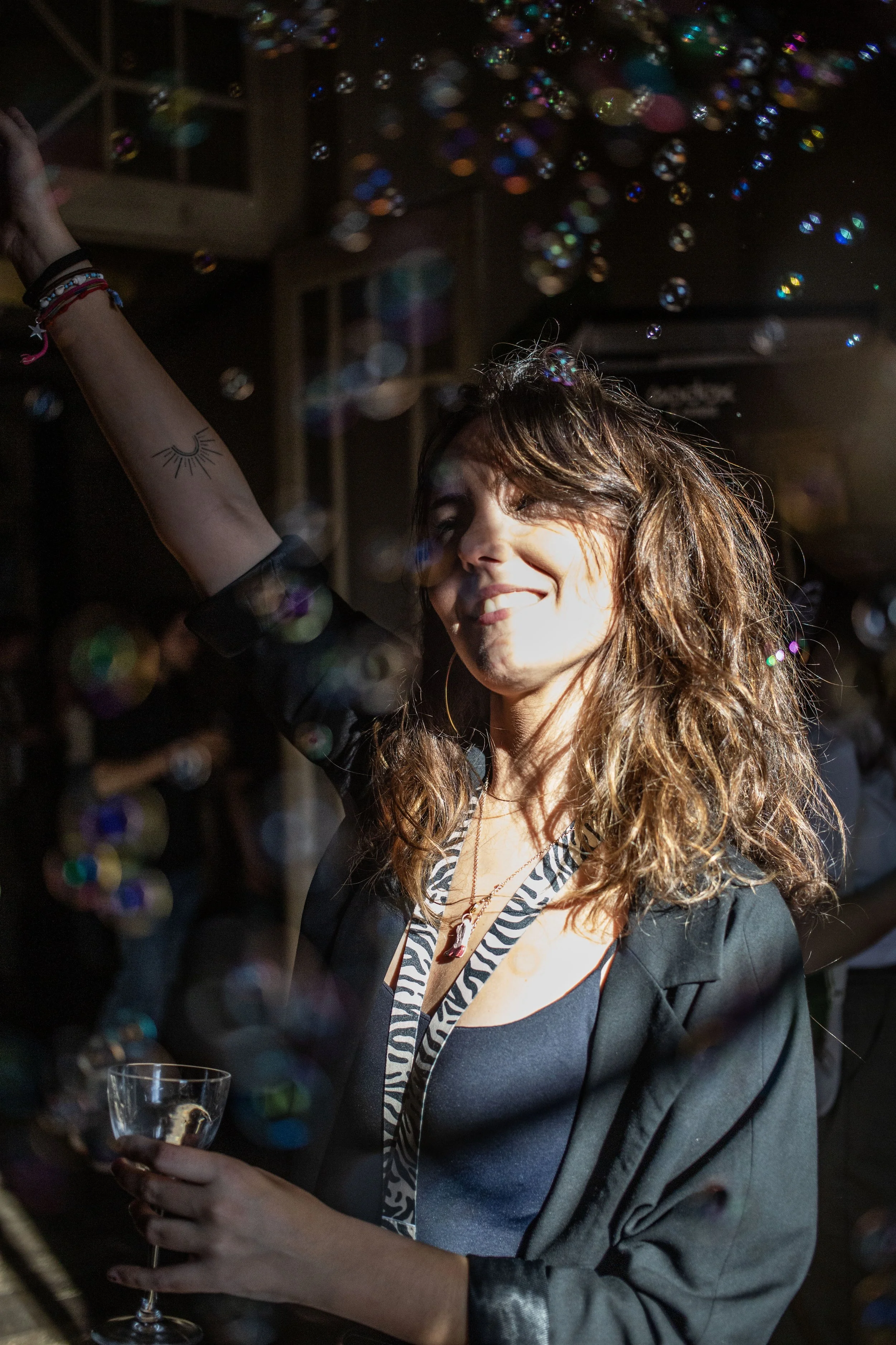 Mujer con cabello rizado y camiseta negra sostiene una copa de vino en una fiesta, rodeada de burbujas de jabón y con expresión alegre. Contenido de redes sociales.