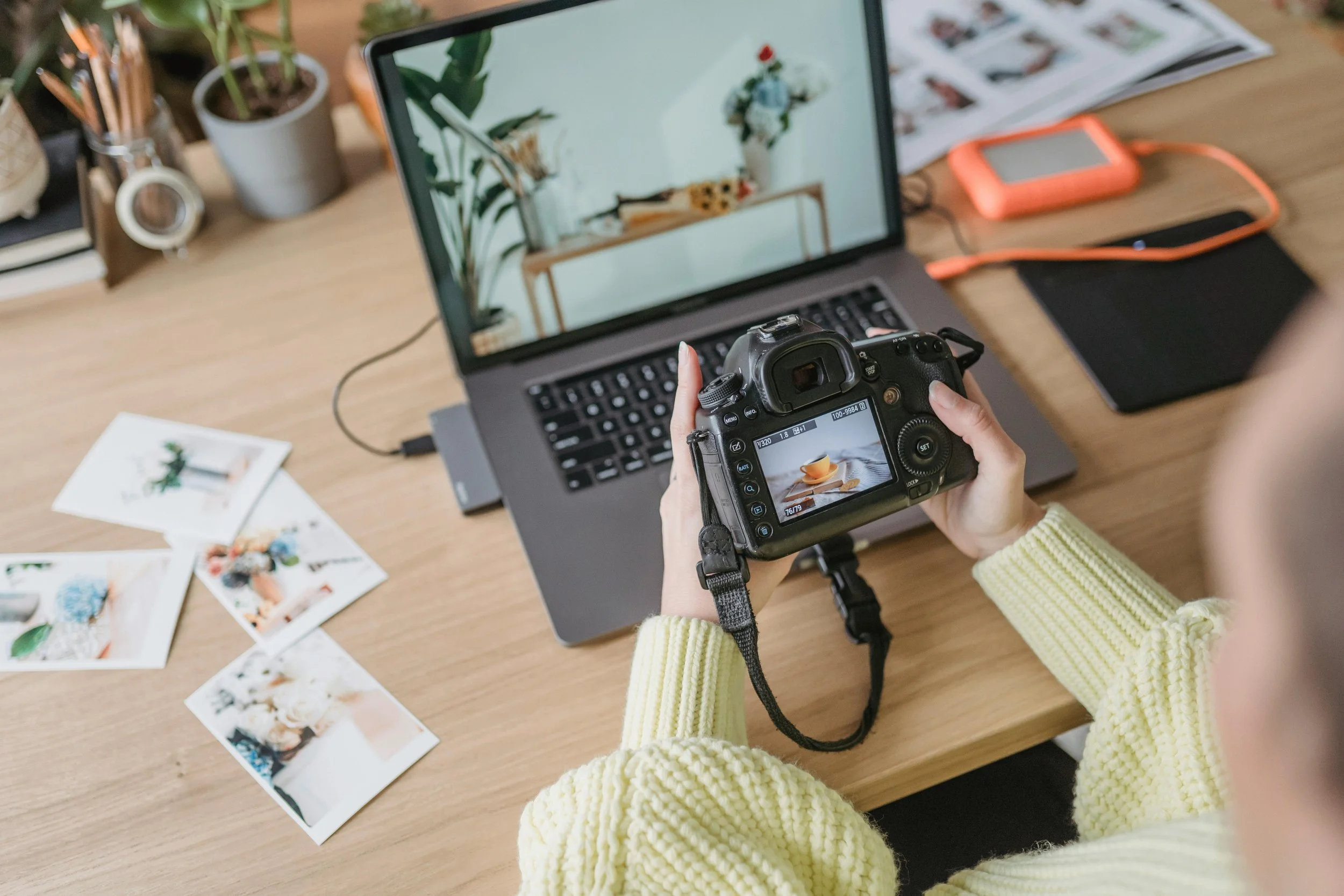 Persona revisando una fotografía en una cámara digital, en un escritorio con varias fotos impresas, plantas y accesorios. Gestión de contenido de redes sociales.