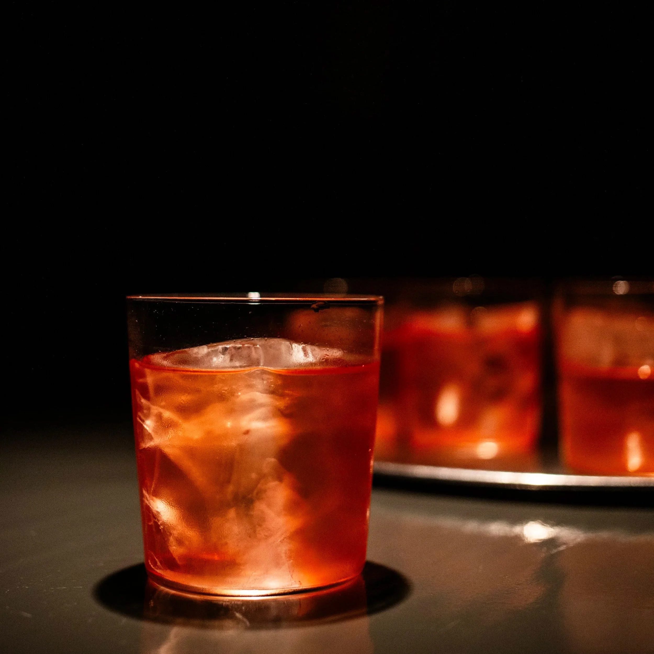 Un vaso con una bebida rosada y hielo, en primer plano, y varios vasos similares en un plato en el fondo, en un ambiente oscuro. Contenido de redes sociales.