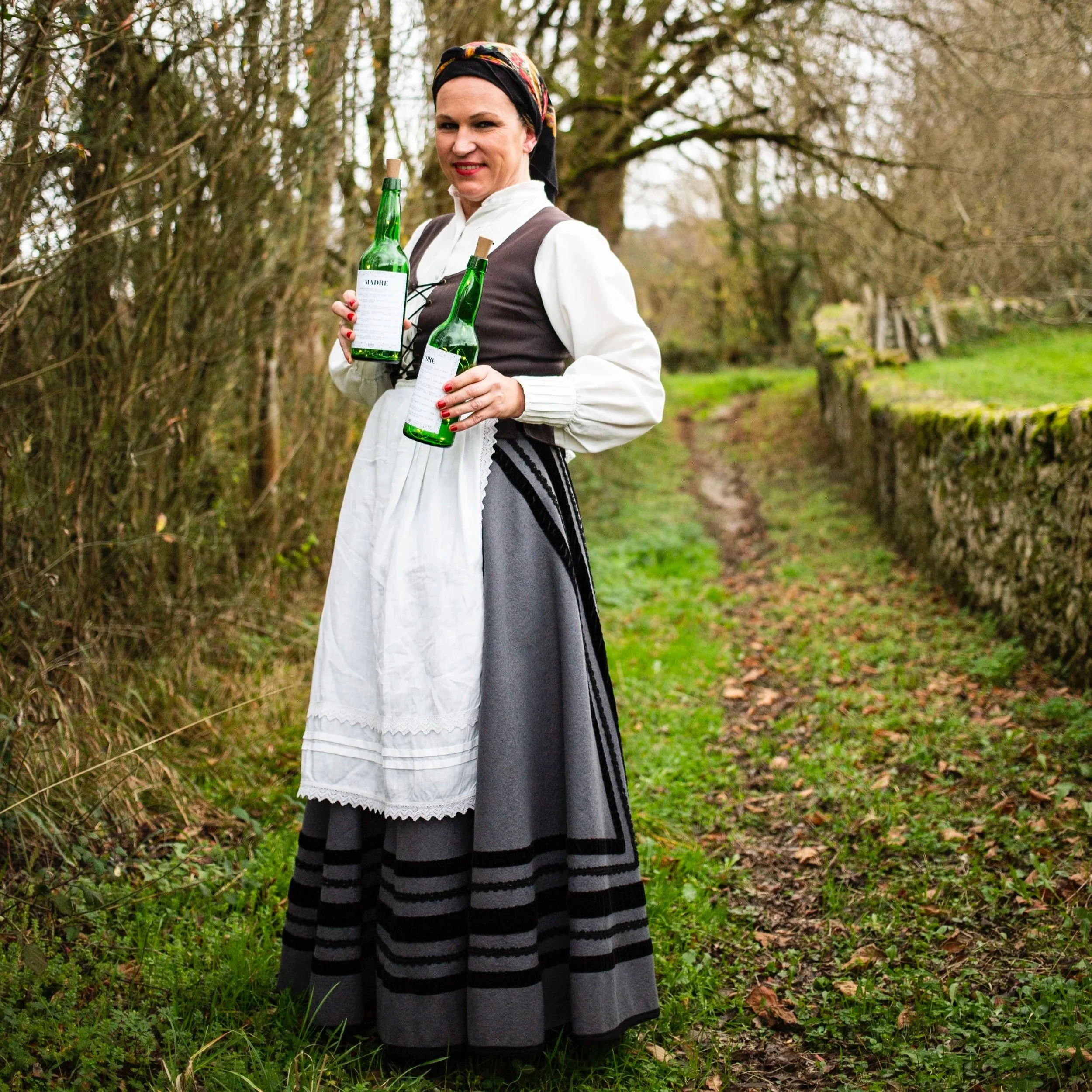 Mujer vestida con ropa tradicional, sosteniendo dos botellas de bebida en un camino de tierra rodeado de árboles y un muro de piedra. Contenido de redes sociales.
