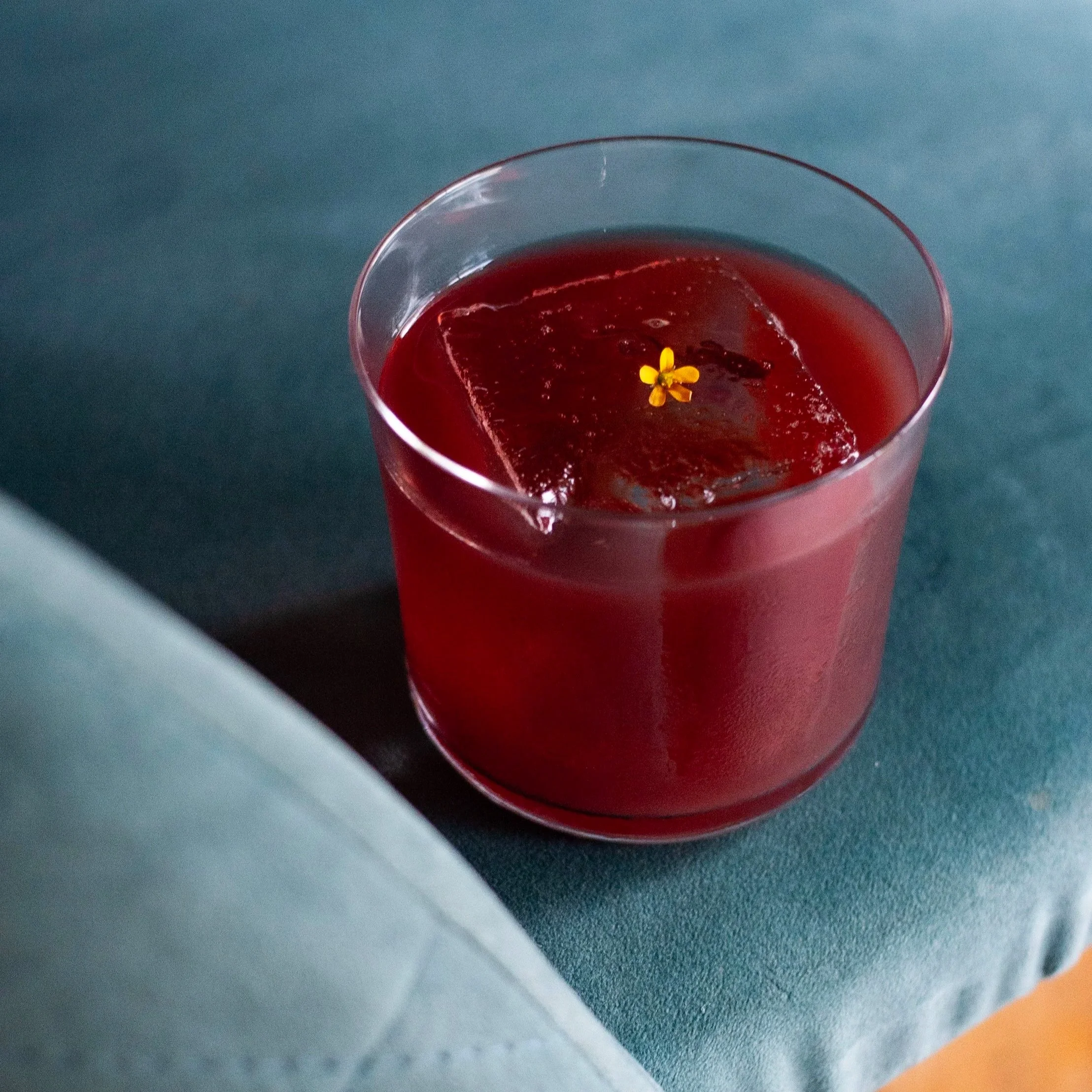 Vaso de vidrio con bebida roja y hielo, decorado con una pequeña flor amarilla en la superficie. Contenido de redes sociales.