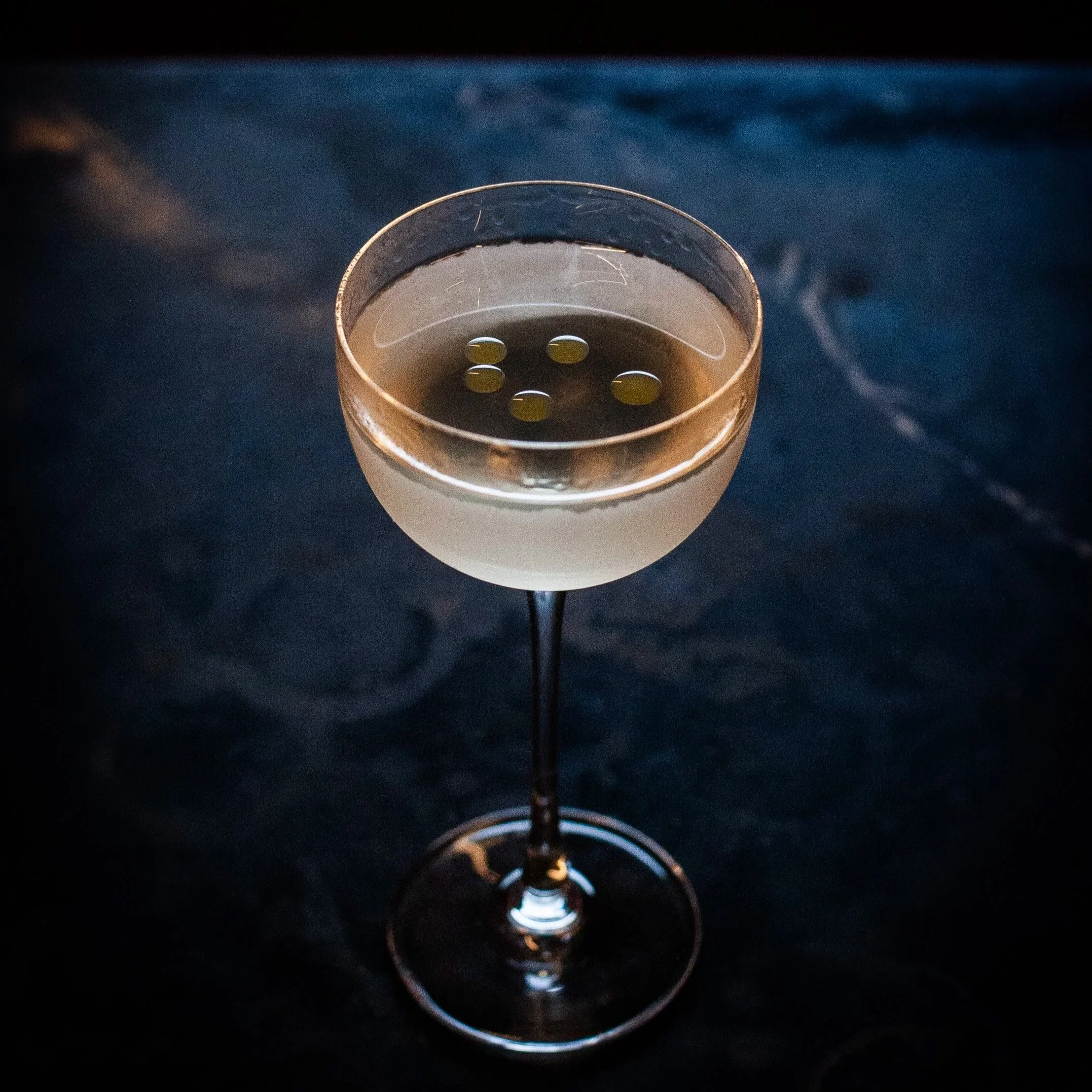 Copa de cóctel con líquido claro y cinco aceitunas verdes en la superficie, sobre una mesa oscura. Contenido de redes sociales.