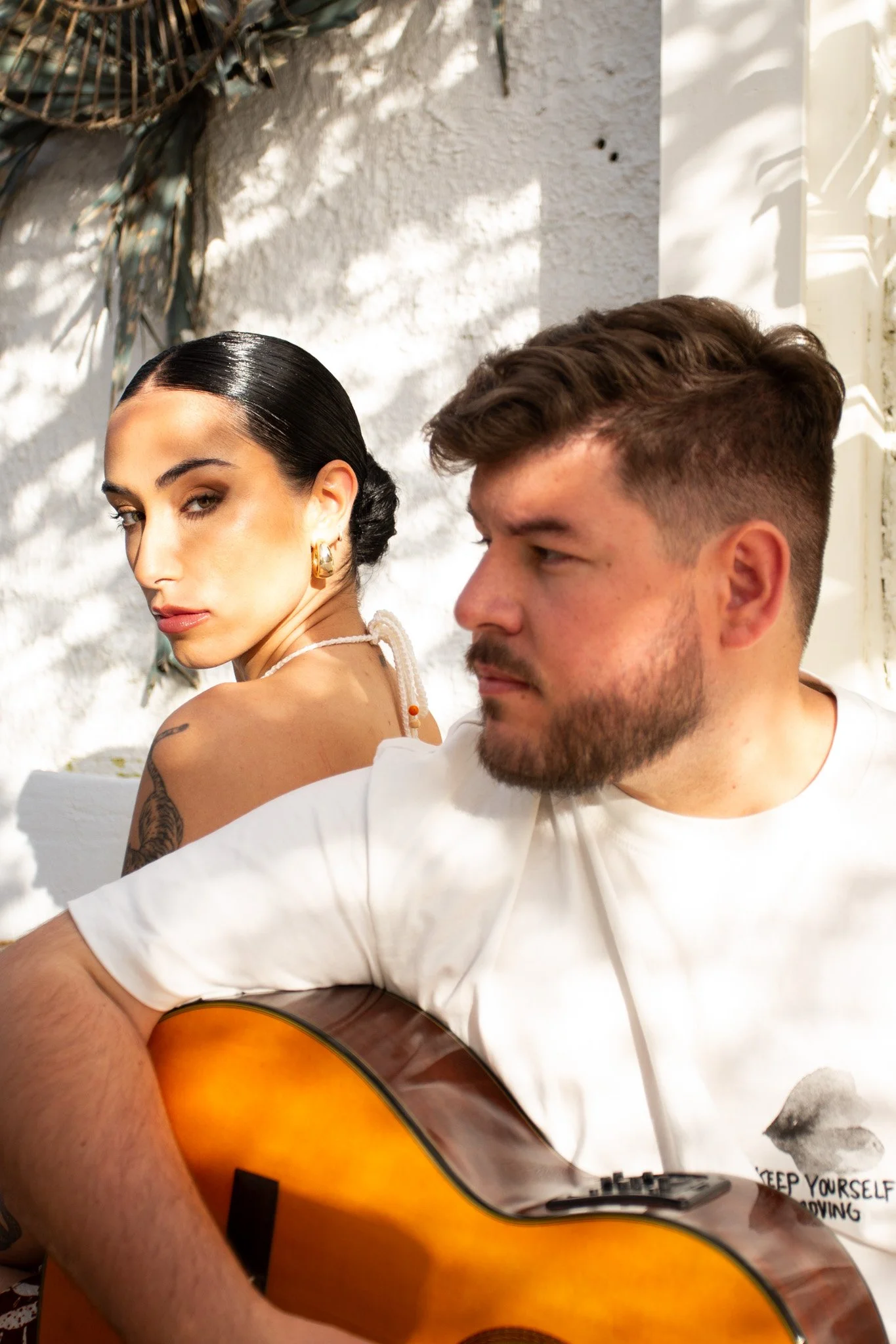 Un hombre tocando guitarra y una mujer sentados en un entorno con luz natural y sombra de plantas en la pared blanca. Contenido de redes sociales.