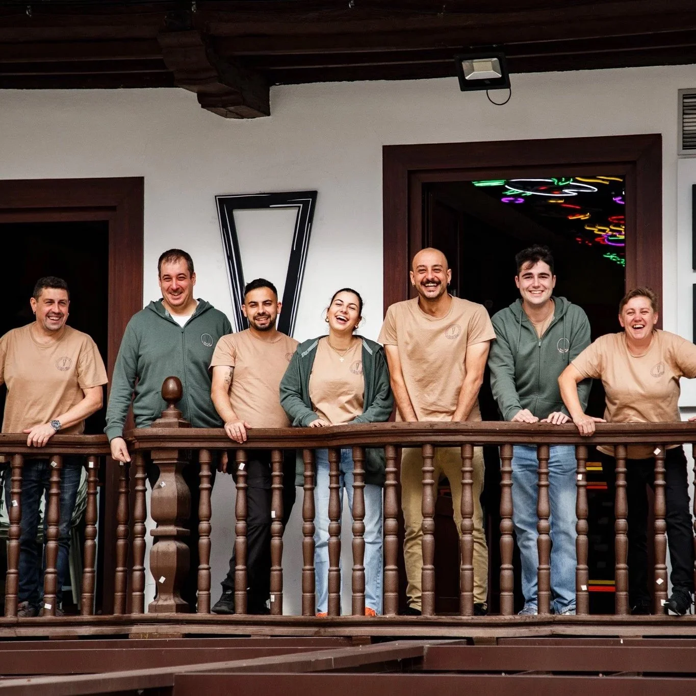 Grupo de siete personas sonriendo en una terraza de interior, algunos con camisetas beige y otros con sudaderas verdes, frente a una barandilla de madera. Contenido de redes sociales.
