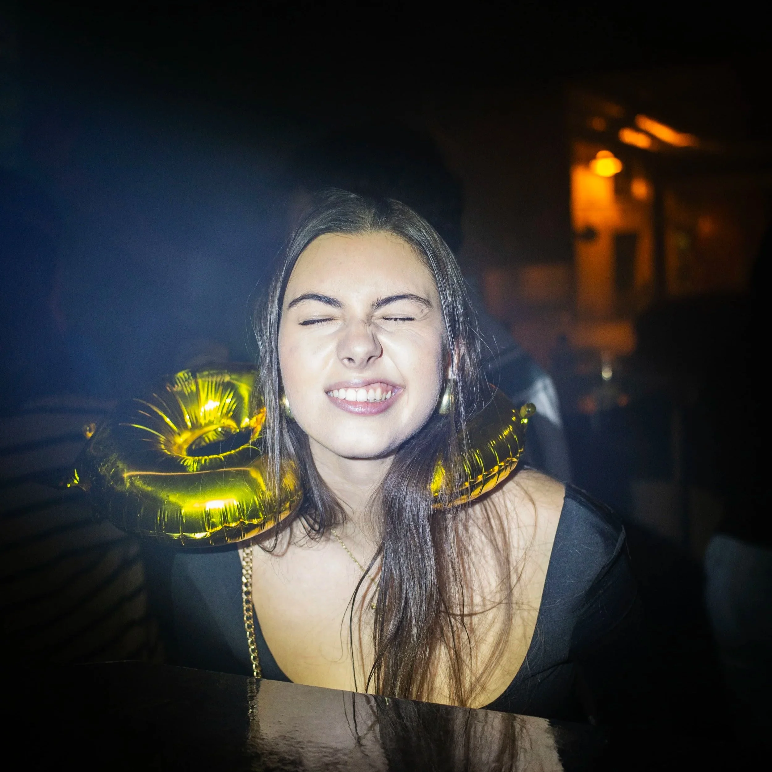 Mujer con expresión de alegría en una fiesta, lleva globos dorados en forma de anillo alrededor del cuello. Contenido de redes sociales.