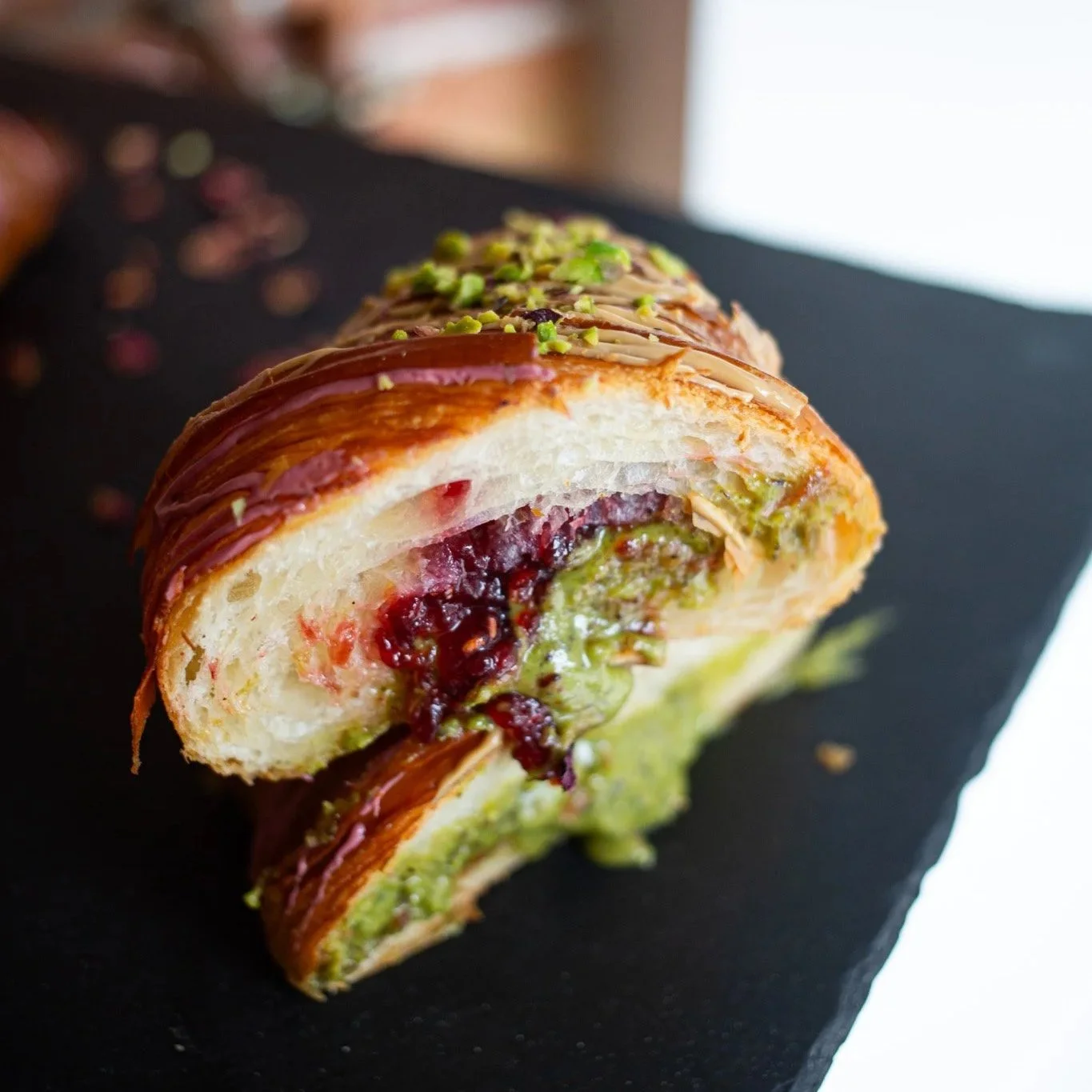 Rebanada de pastel con relleno de mermelada de frutas y relleno de crema, cubierto con glaseado de chocolate y decorado con pistachos picados. Contenido de redes sociales.