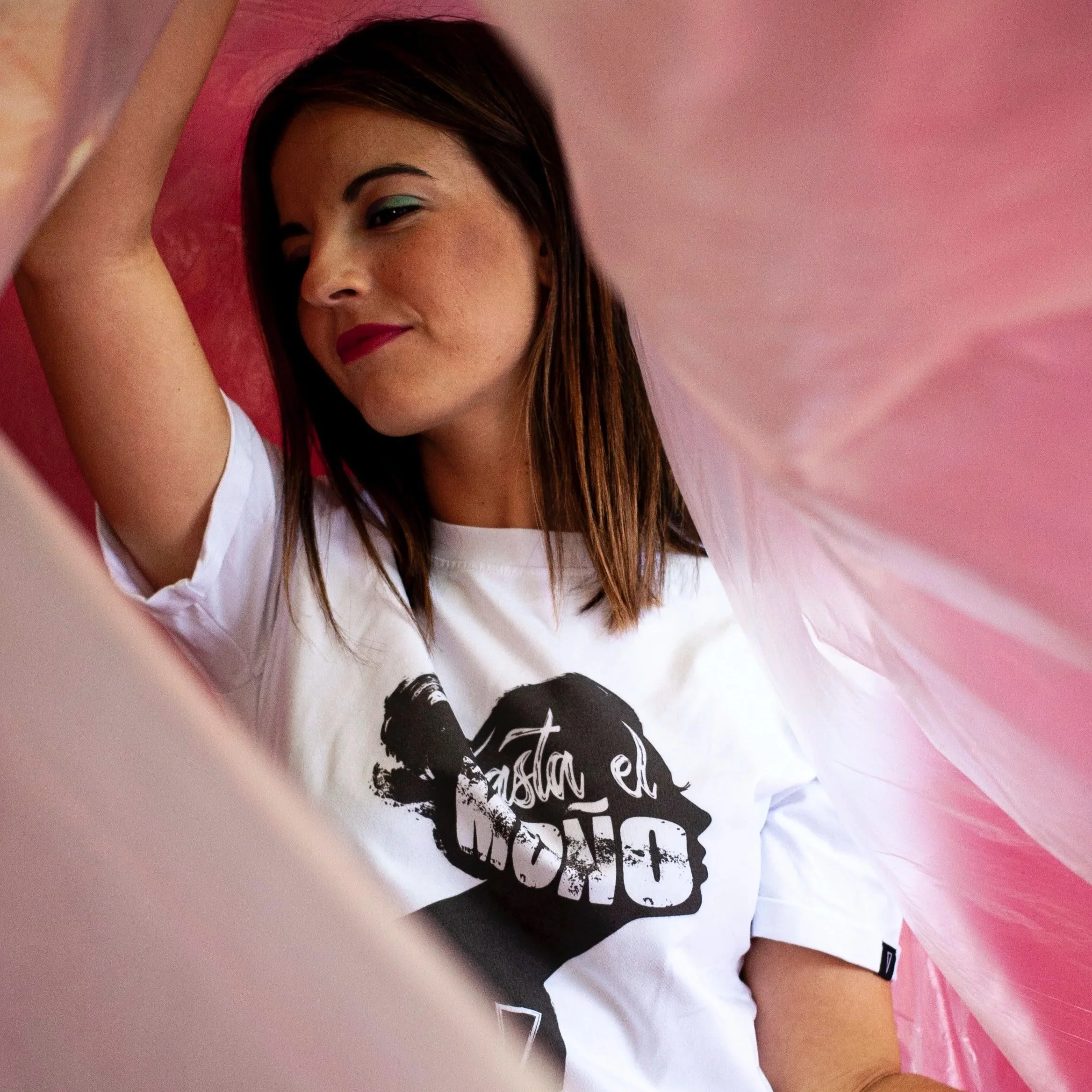 Joven mujer con maquillaje de labios rojos y sombra azul, vistiendo camiseta blanca con texto en negro, posando entre cortinas rosas. Contenido de redes sociales.