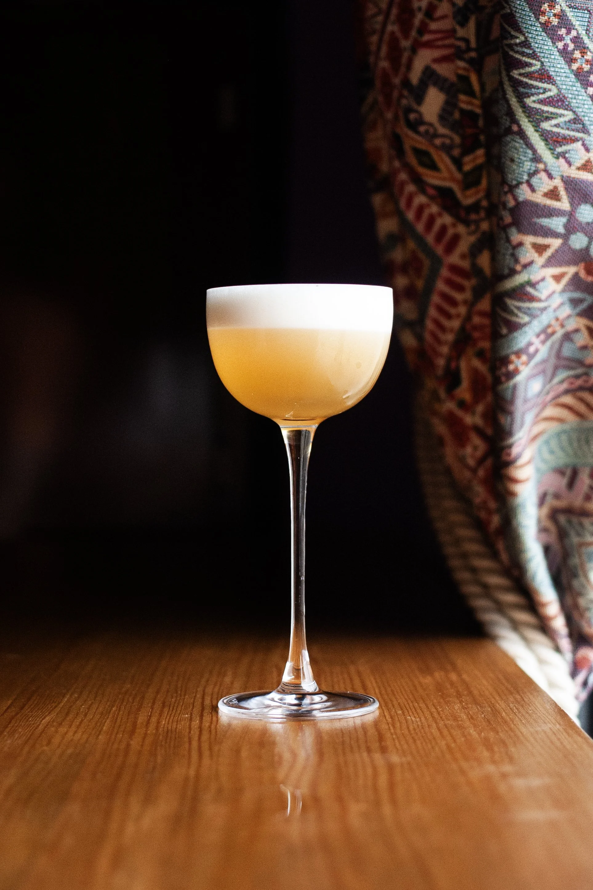 Copa de cóctel con espuma en la parte superior sobre mesa de madera, fondo oscuro y cortina con varios patrones coloridos a la derecha. Contenido de redes sociales.