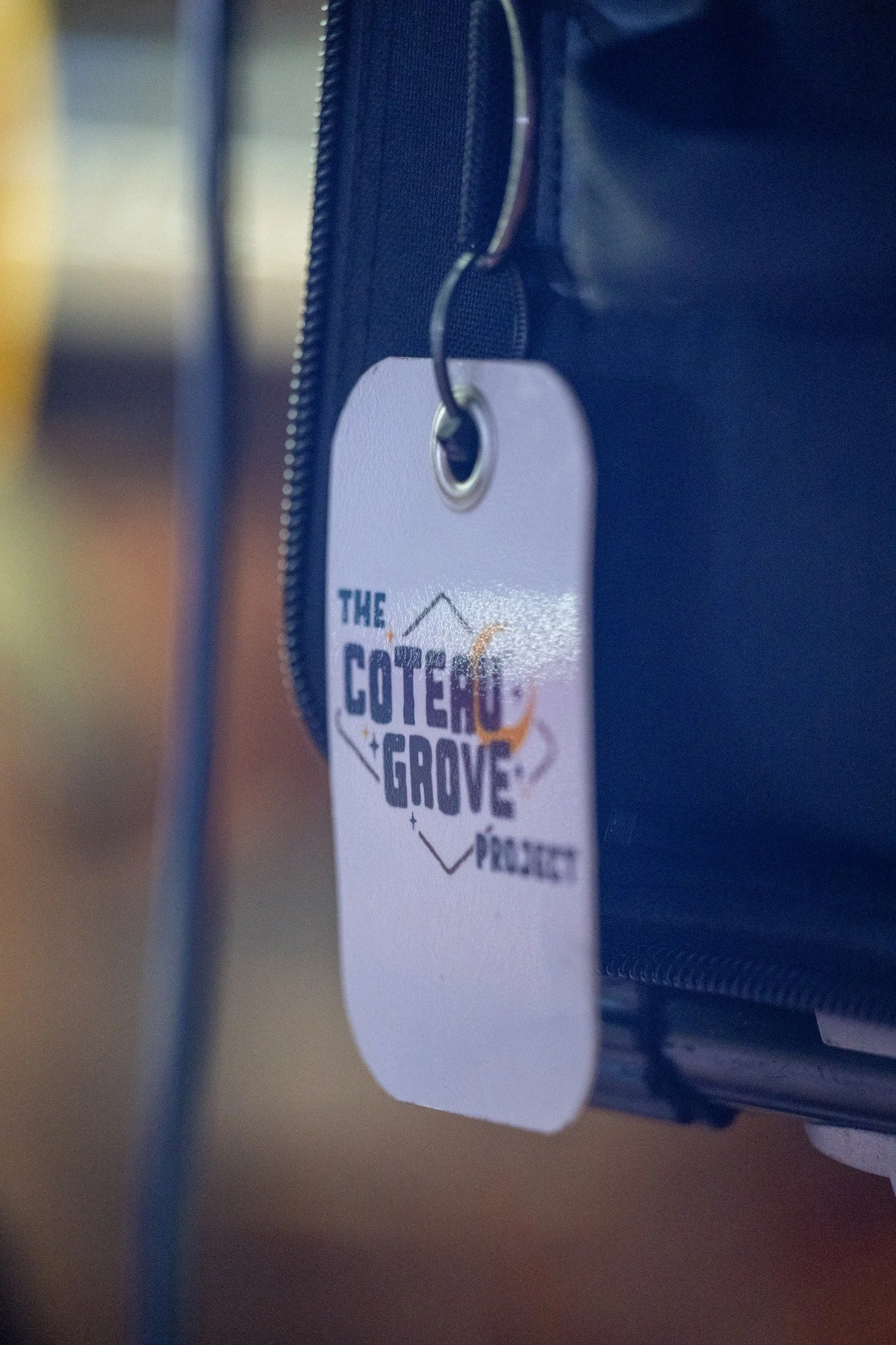 A close-up of a white tag hanging from a zipper on a blue bag, with the logo of The Cotero Grove Project.