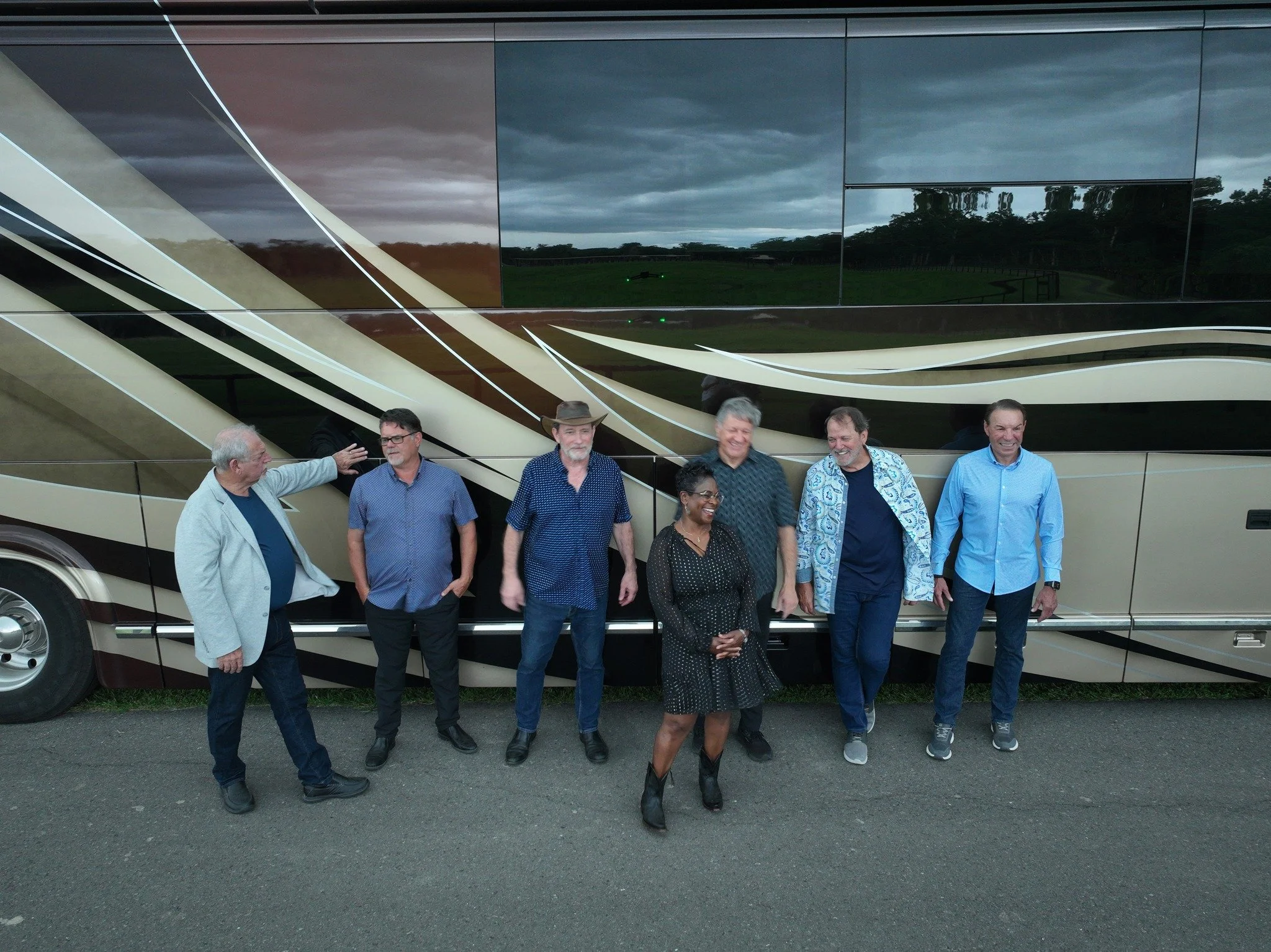 Group of seven people standing in front of a large boat with a reflective surface and abstract patterns, some smiling and talking, outdoors