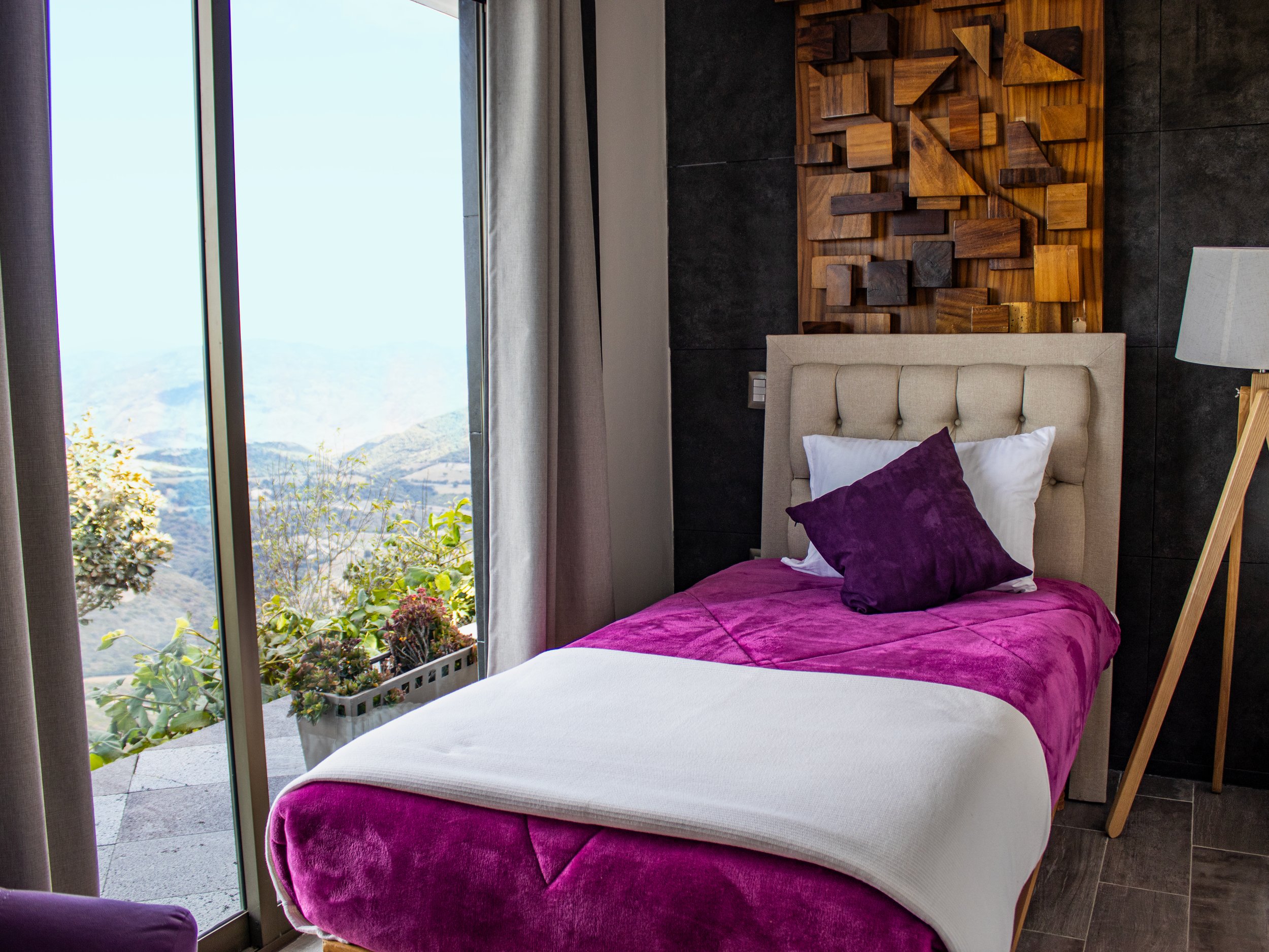 A cozy bedroom with a bed that has a tufted beige headboard, purple bedding, and a purple pillow. There is a wooden wall art behind the bed and a white lamp on a wooden tripod next to it. A sliding glass door on the left opens to a balcony with potted plants, revealing a mountain landscape outside.
