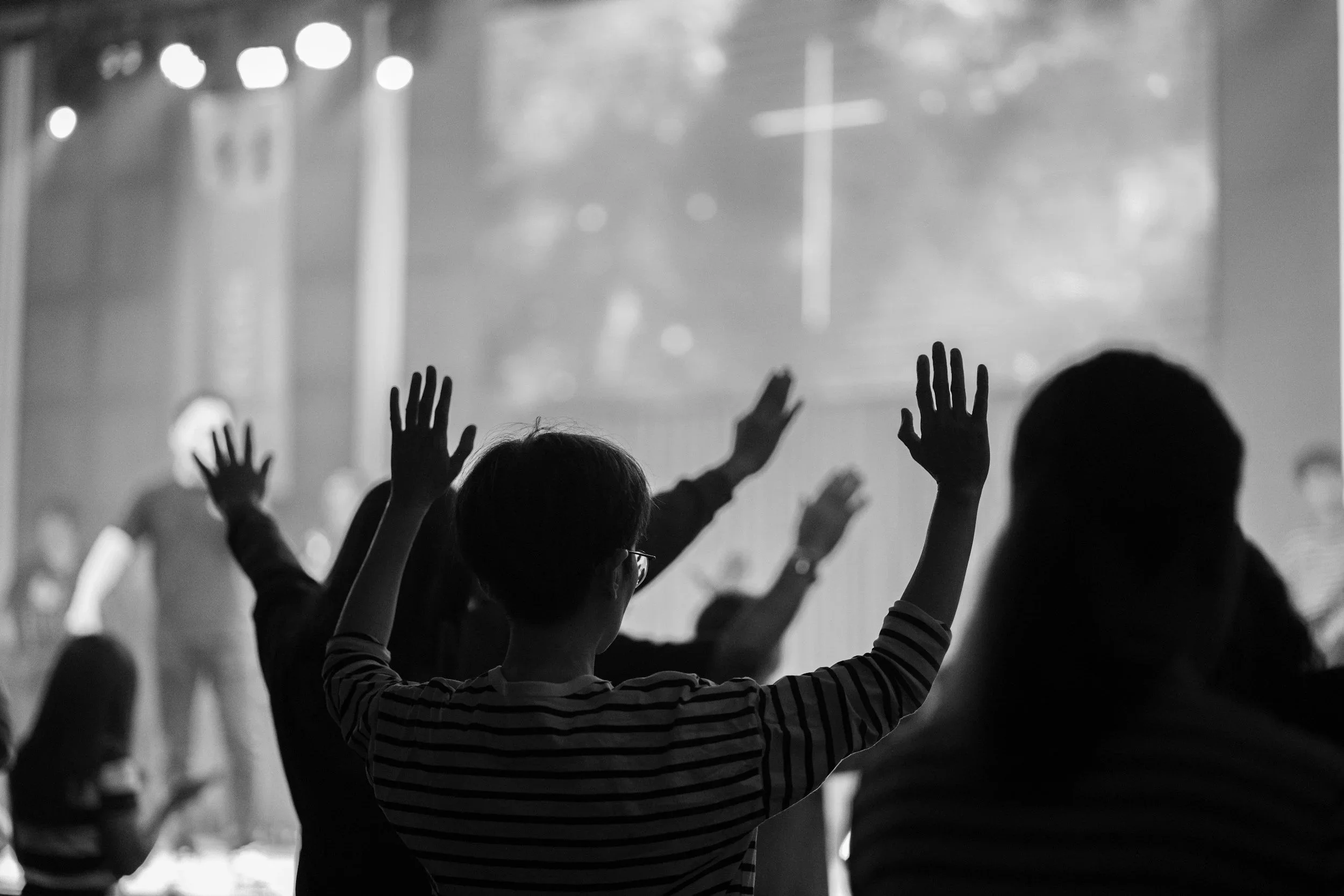 People raising their hands in a dark, crowded setting, possibly at a concert or worship service, with stage lights and blurred background.