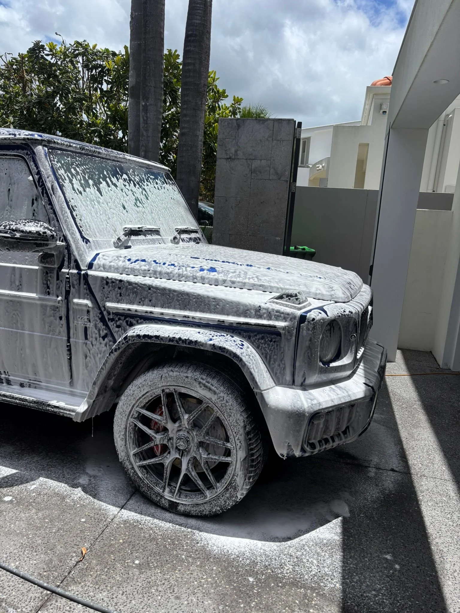 New black SUV covered in soap suds during car wash, parked outside modern house with white walls and palm trees in background.