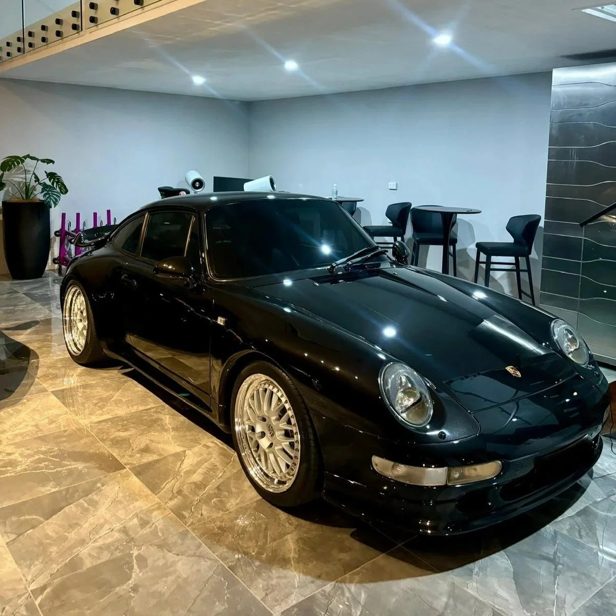 Black Porsche 911 sports car parked indoors on a marble floor, with a potted plant and bar stools in the background.