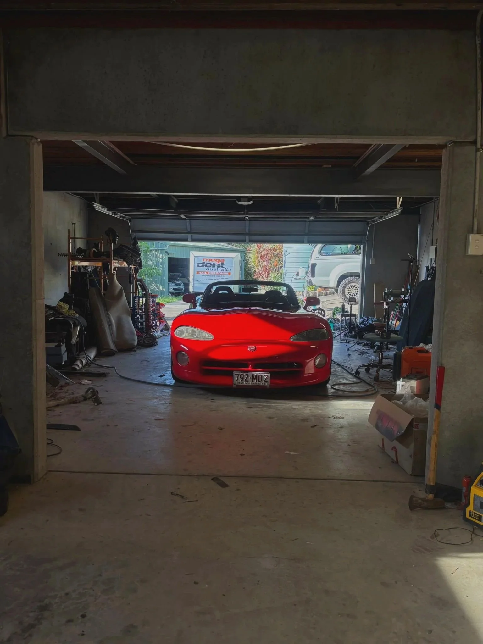 A red sports car parked inside a garage with an open garage door. The garage has various tools and equipment scattered around, and some vehicles are visible outside.