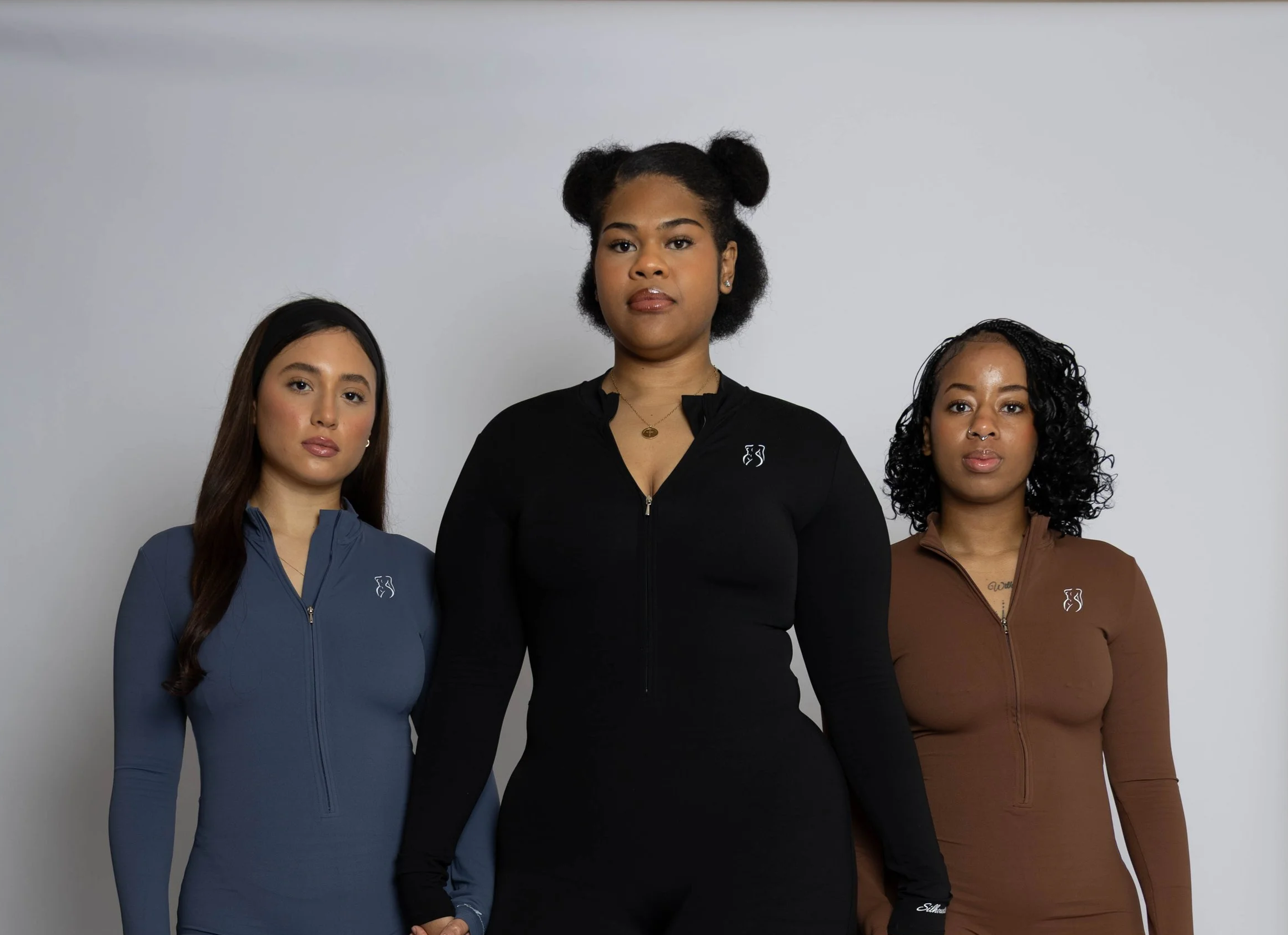 Three women standing side by side against a gray background, wearing athletic zip-up jackets in blue, black, and brown, with hair styled in different ways, and looking directly at the camera.
