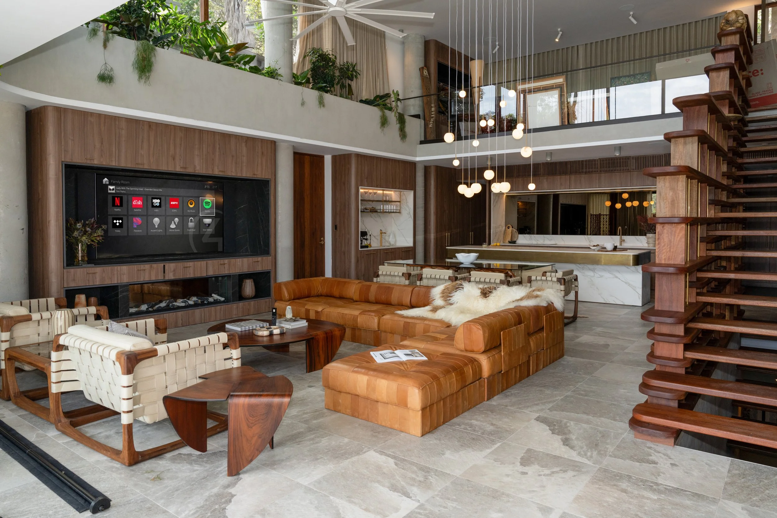 Modern living room with leather sectional sofa, striped armchairs, wood coffee tables, a large flat-screen TV on a wooden wall, a staircase to the right, and a high ceiling with a white fan and hanging globe lights.