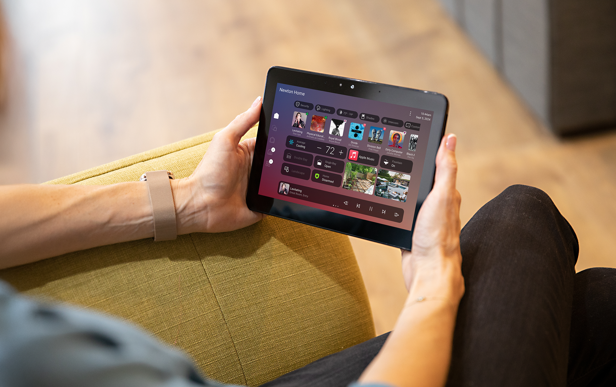 Person holding a tablet displaying a smart home control app. The app shows various controls and media options for a smart home, with a background of a yellow couch and wooden floor.