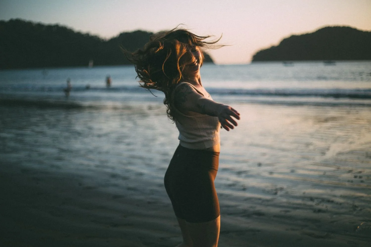 A woman with long hair standing at the edge of the water on a beach during sunset, with her arms outstretched and hair blowing in the wind.