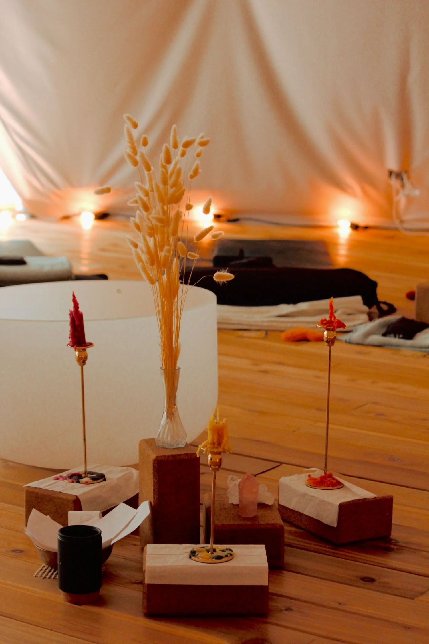 Decorative arrangement with dried flowers, candles, and small objects on wooden blocks, in a cozy indoor setting with soft lighting and blankets in the background.