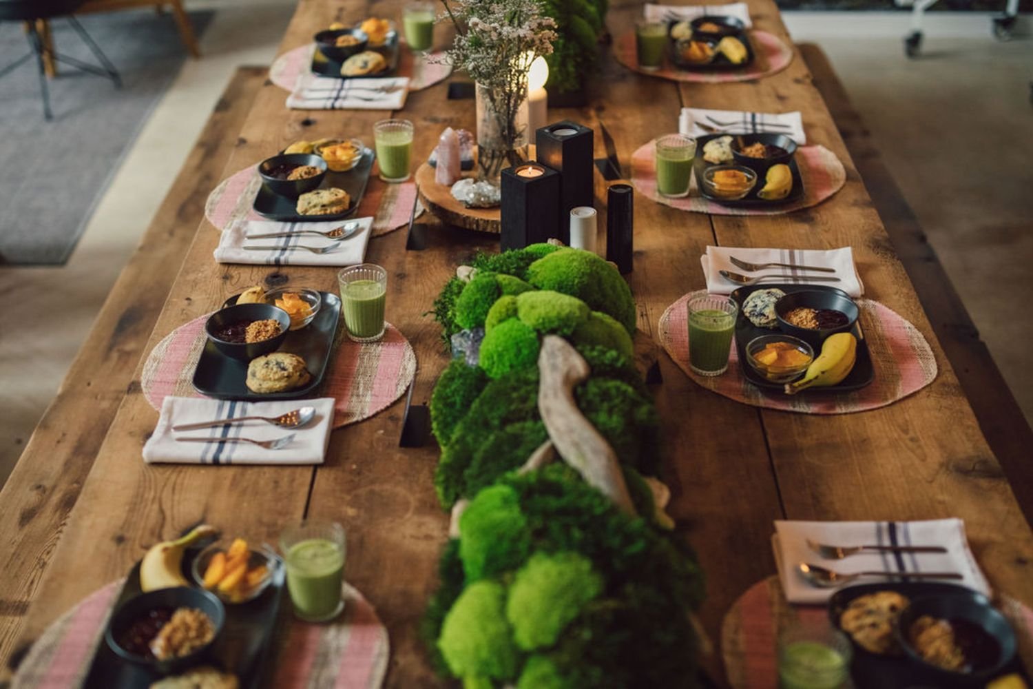 A rustic wooden dining table set for a meal with six place settings. Each setting includes a black tray with bowls of soup or stew, a cookie, and a small dish of fruit. Green smoothies are placed next to each setting. The table is decorated with a central arrangement of a vase of flowers, candles, and decorative stones, creating a cozy and natural ambiance.