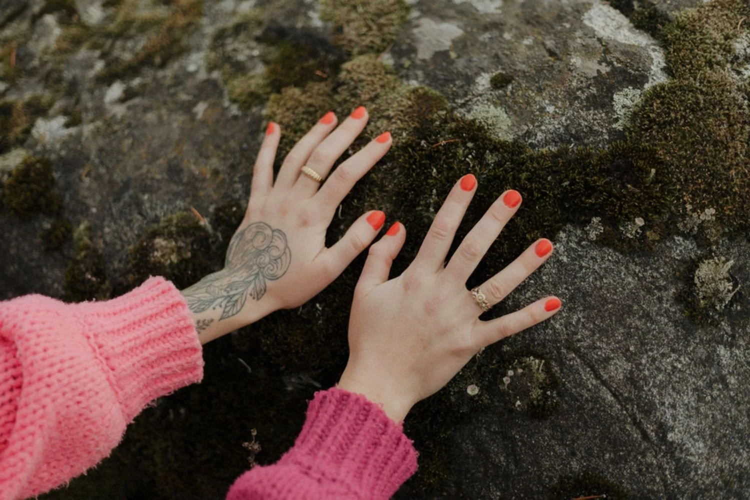 Two hands with orange nail polish placed on a moss-covered rock surface, one hand has a tattoo and wears rings, and both wear pink knitted sweaters.
