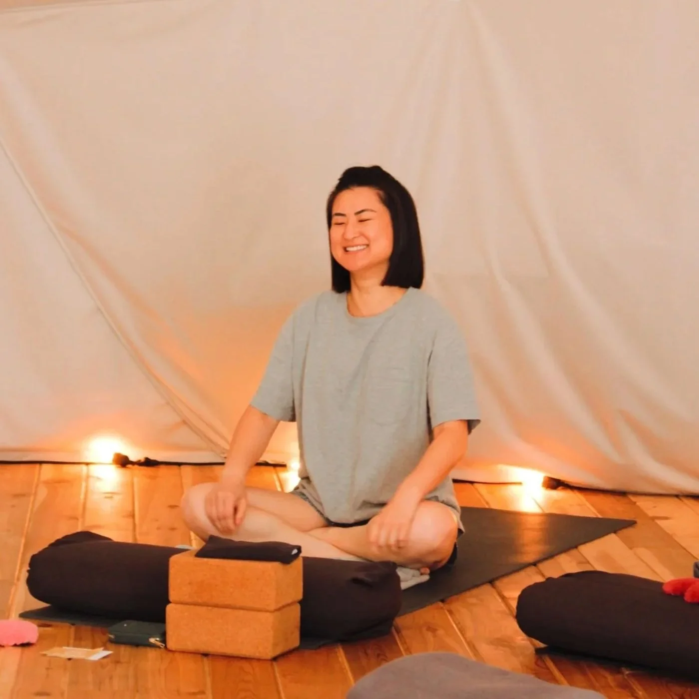 Woman smiling with eyes closed, sitting cross-legged on a yoga mat in a cozy, softly lit room with wood flooring and cream-colored drapes.
