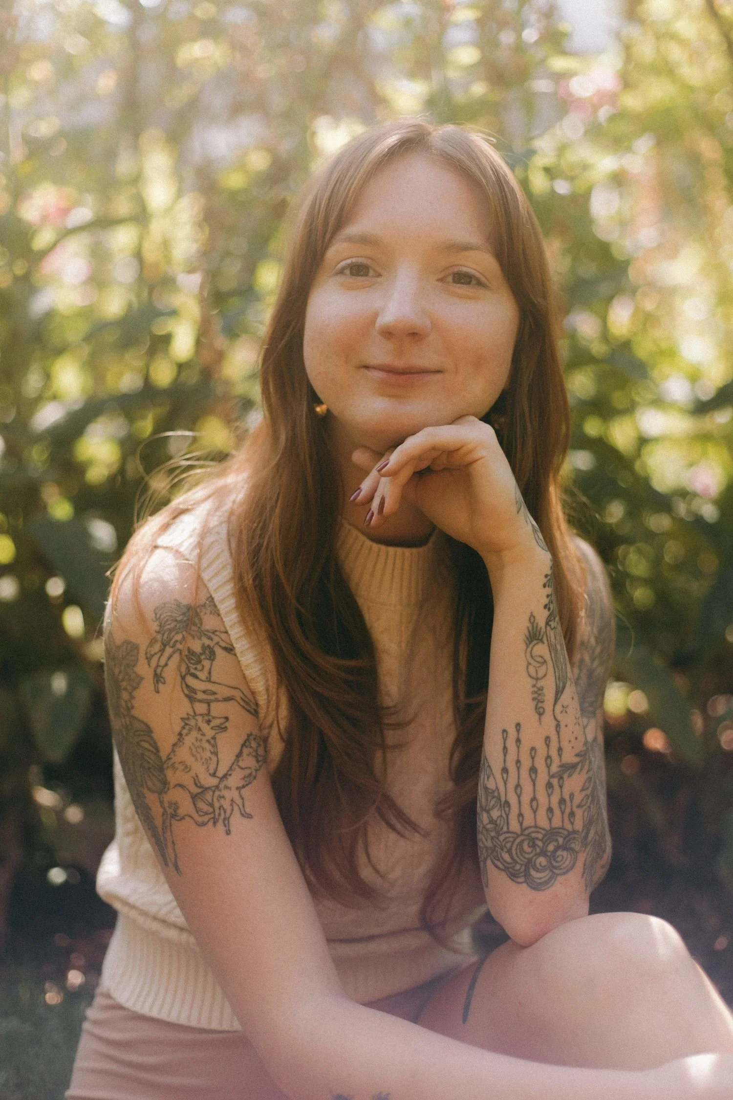 Woman with long hair and tattoos on her arms, sitting outdoors with a background of green foliage and sunlight filtering through trees.