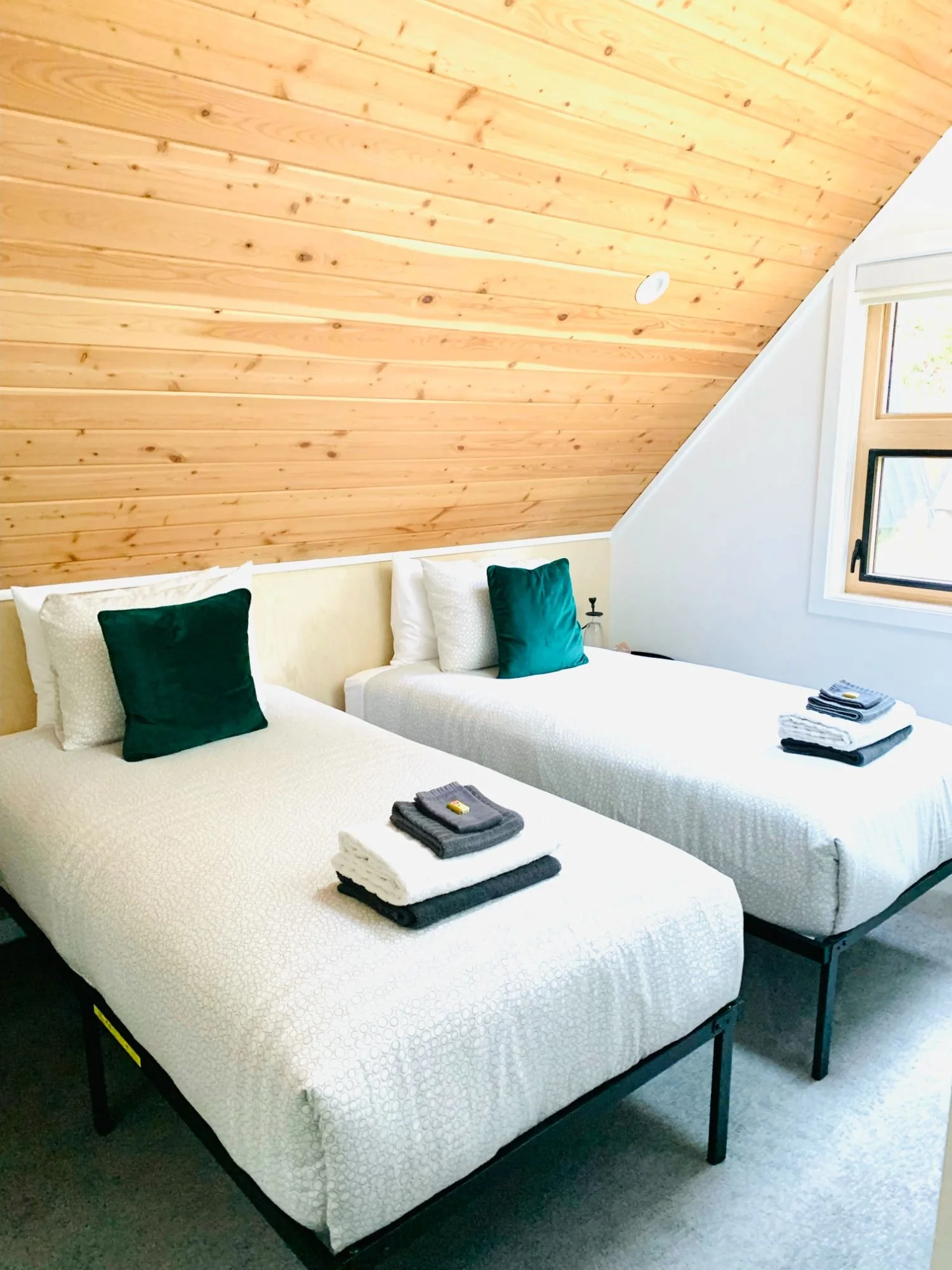 Two neatly made beds with white bedding and dark green pillows, set in a room with a wooden sloped ceiling and a window on the right side.
