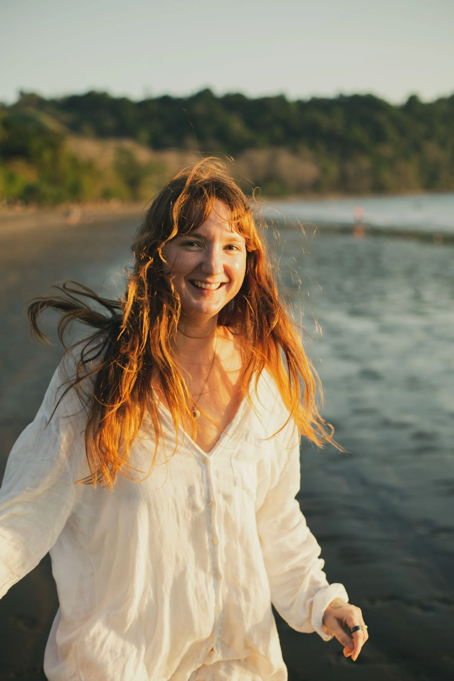Smiling woman with long hair wearing a white shirt, standing on a beach during sunset.