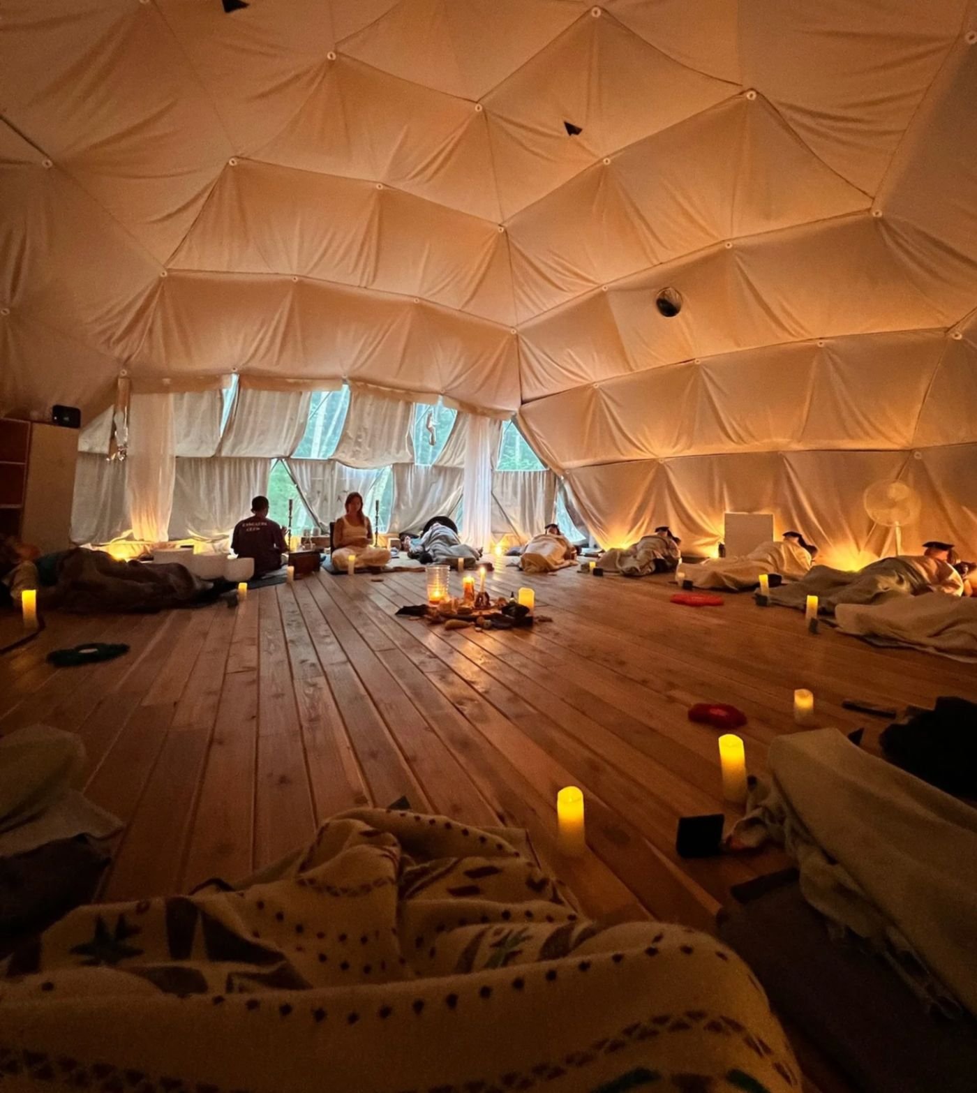 A cozy indoor space with wooden flooring, dim candle lighting, and people lying on their stomachs with heads down, possibly receiving massages. The ceiling is inflatable and tent-like, resembling a geodesic dome, with large windows and curtains along one side. Candles and personal belongings are scattered around, creating a relaxed and tranquil atmosphere.