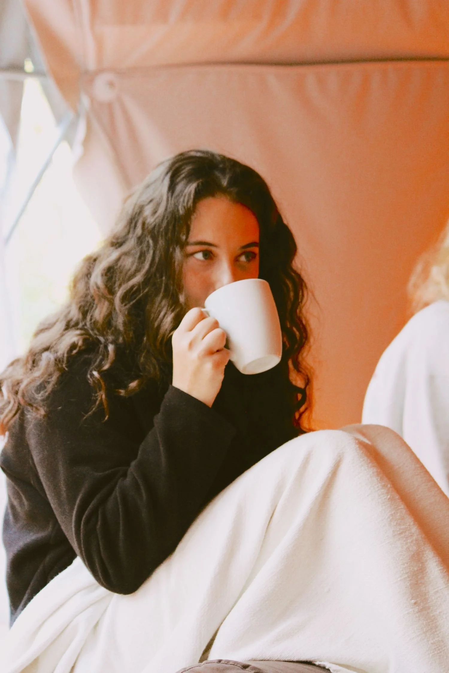 A woman with curly dark hair sitting indoors and drinking from a white mug.