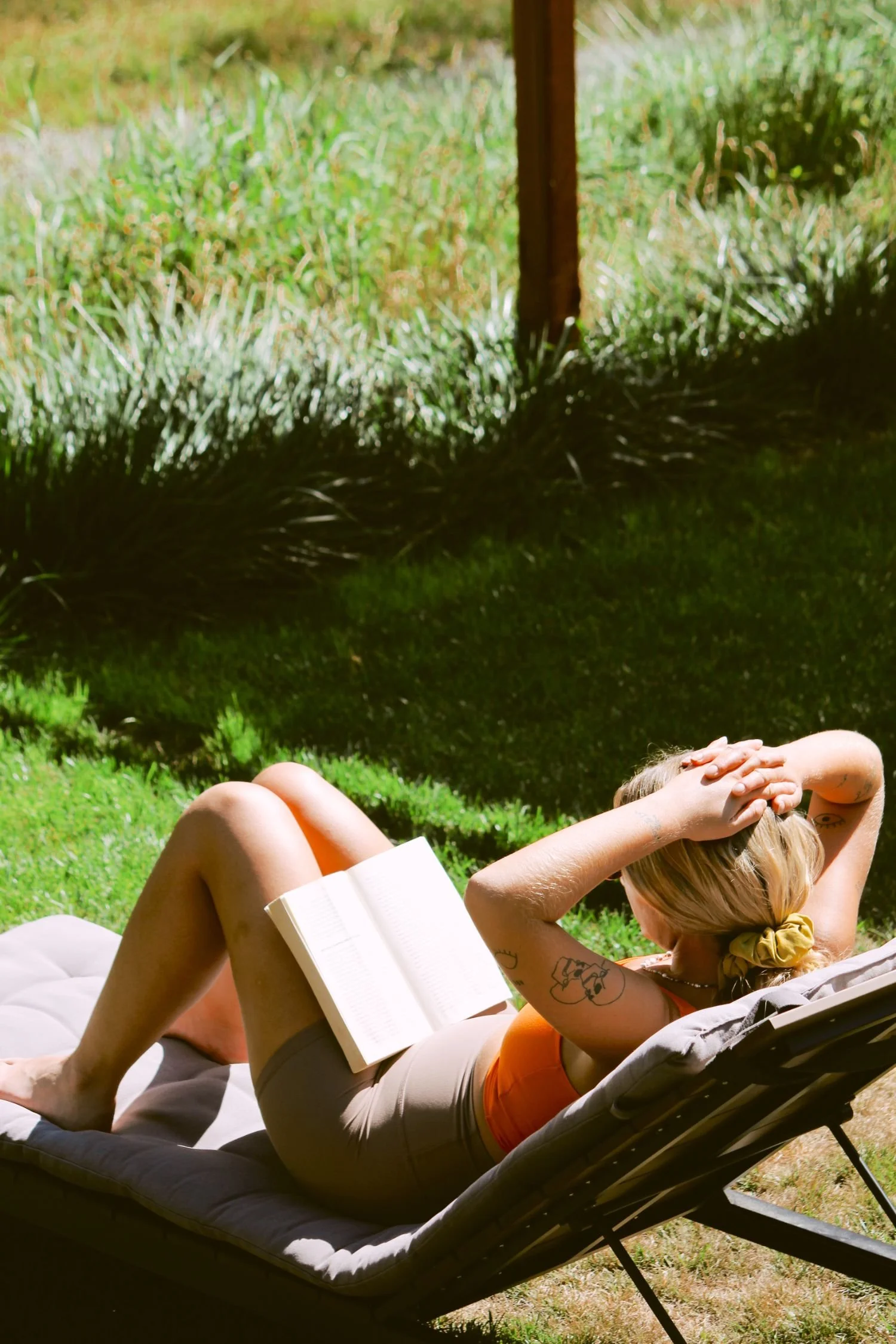 A woman laying on a lounge chair outdoors, reading a book with her hands behind her head and wearing an orange top and beige shorts.