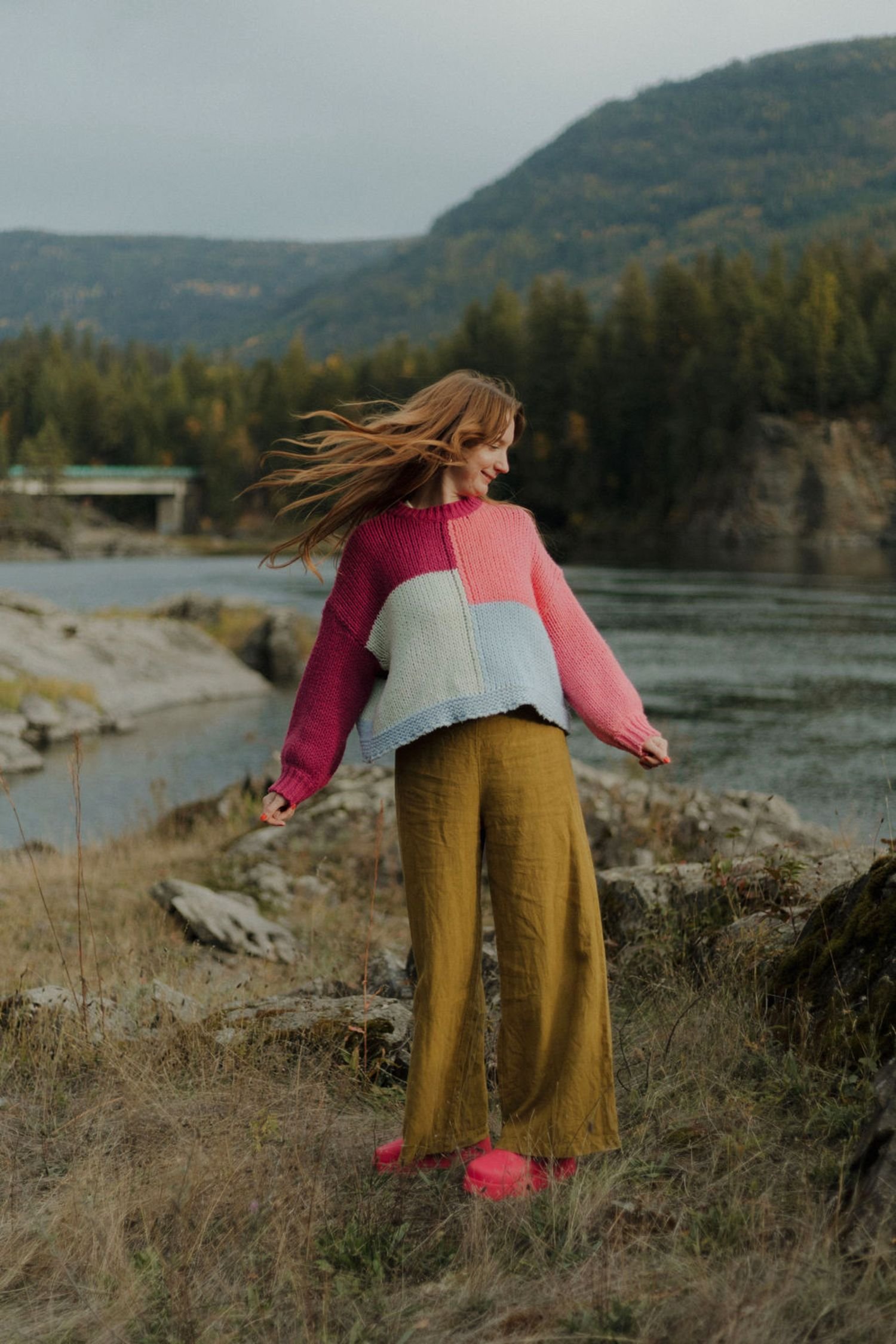 A woman standing on a grassy area by a river, wearing a colorful sweater, yellow pants, and red shoes, with her hair blowing in the wind.