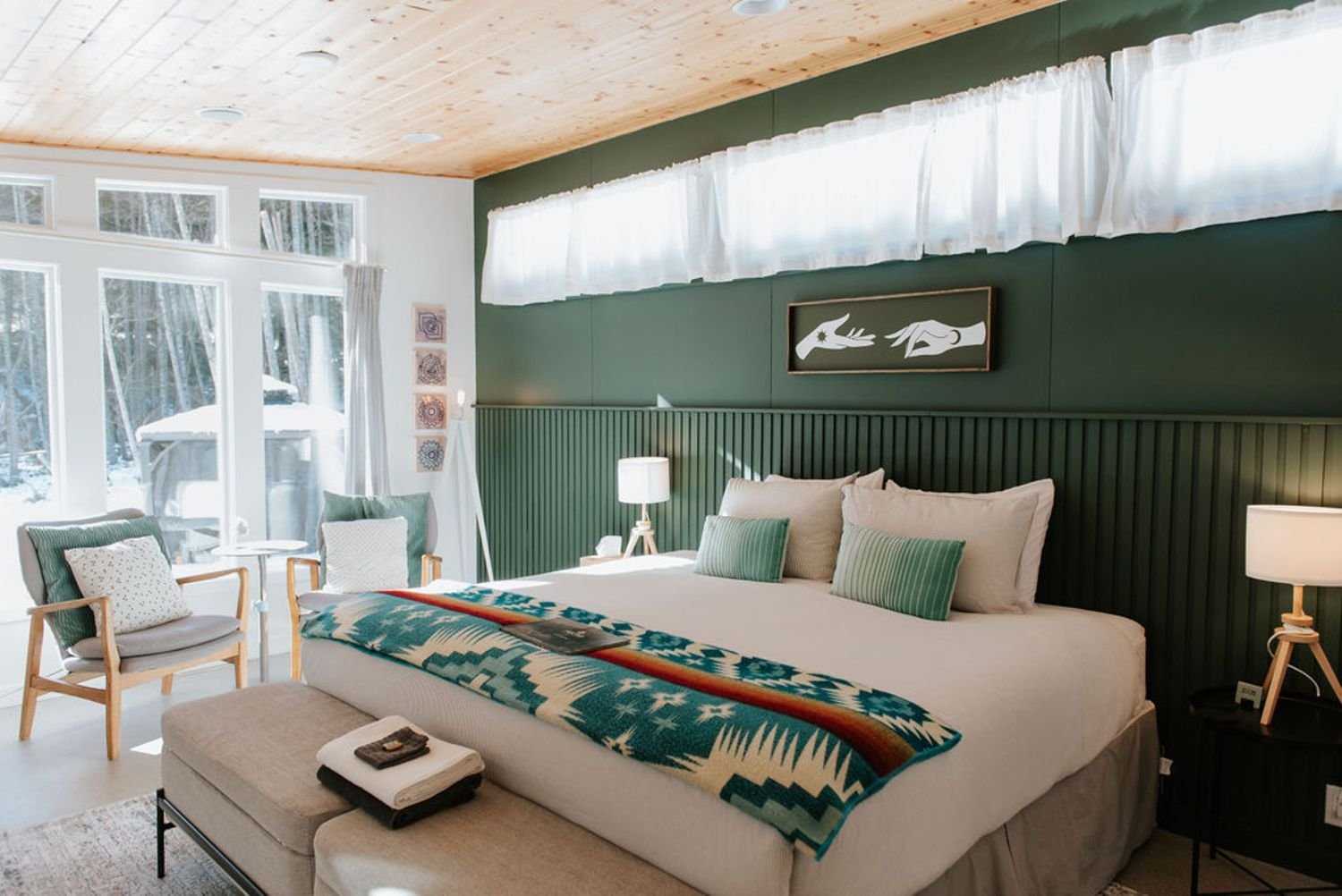 A cozy bedroom with large windows showing a snowy outdoor scene. The room has a green accent wall with artwork, a king-sized bed with pillows and a southwestern-patterned blanket, nightstands with lamps, and a seating area with chairs and a small table.