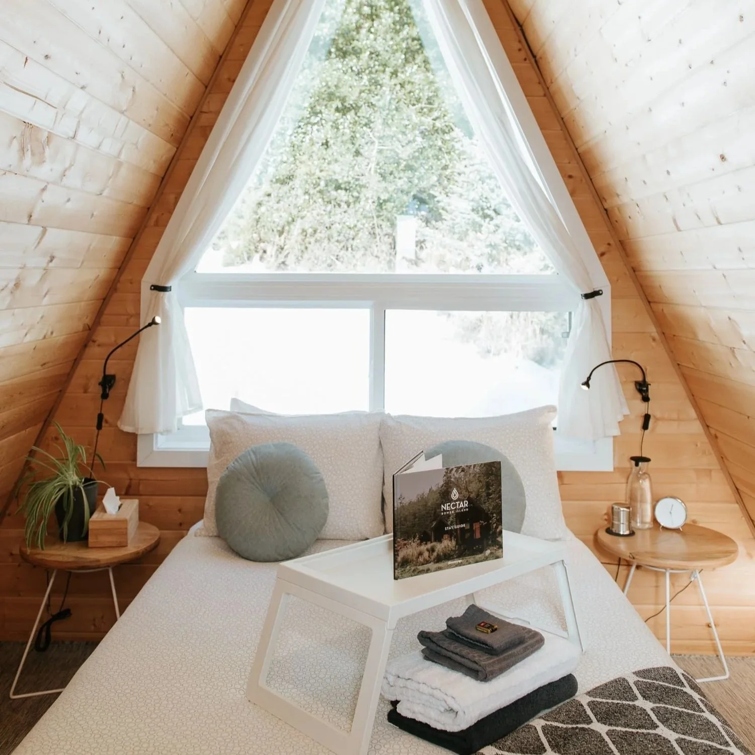 A cozy bedroom with wooden walls and a triangular window showing a tree outside. The bed has white bedding, two round pillows, and a tray with folded towels and a brochure on top. There are two small side tables with lamps and decorative items, including a plant, a clock, and a candle.