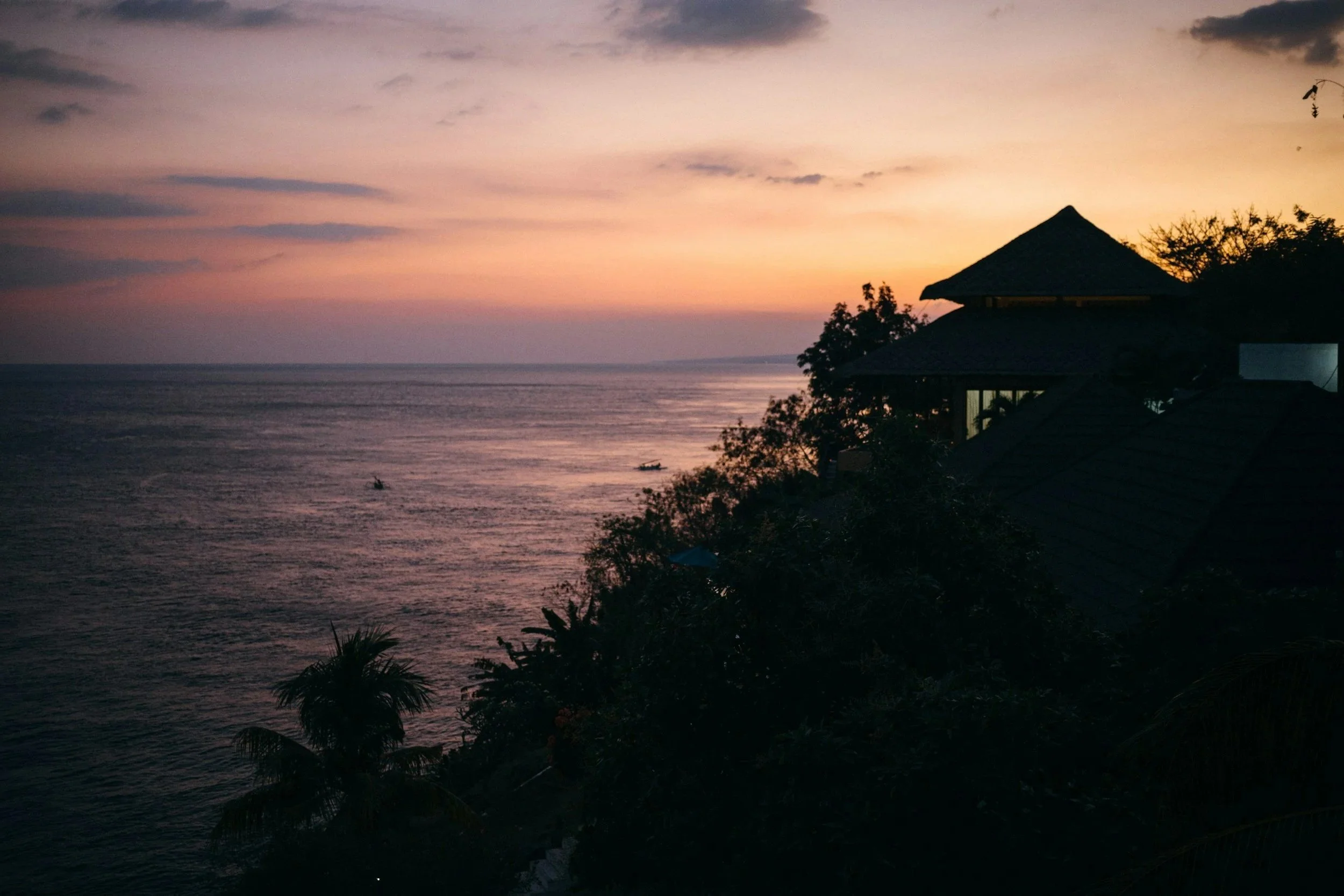 Sunset view over the ocean with a silhouette of a building, trees, and boats on the water.