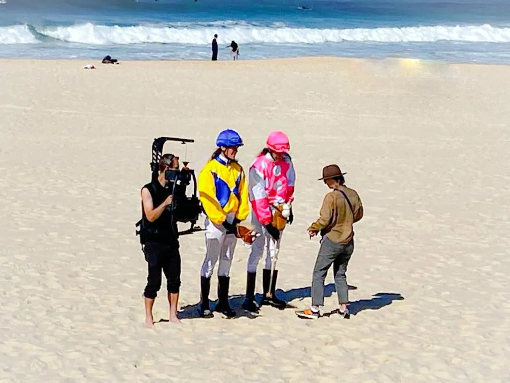 People on a sandy beach, filming a scene with two horses and two actors dressed in colorful costumes, near the ocean.