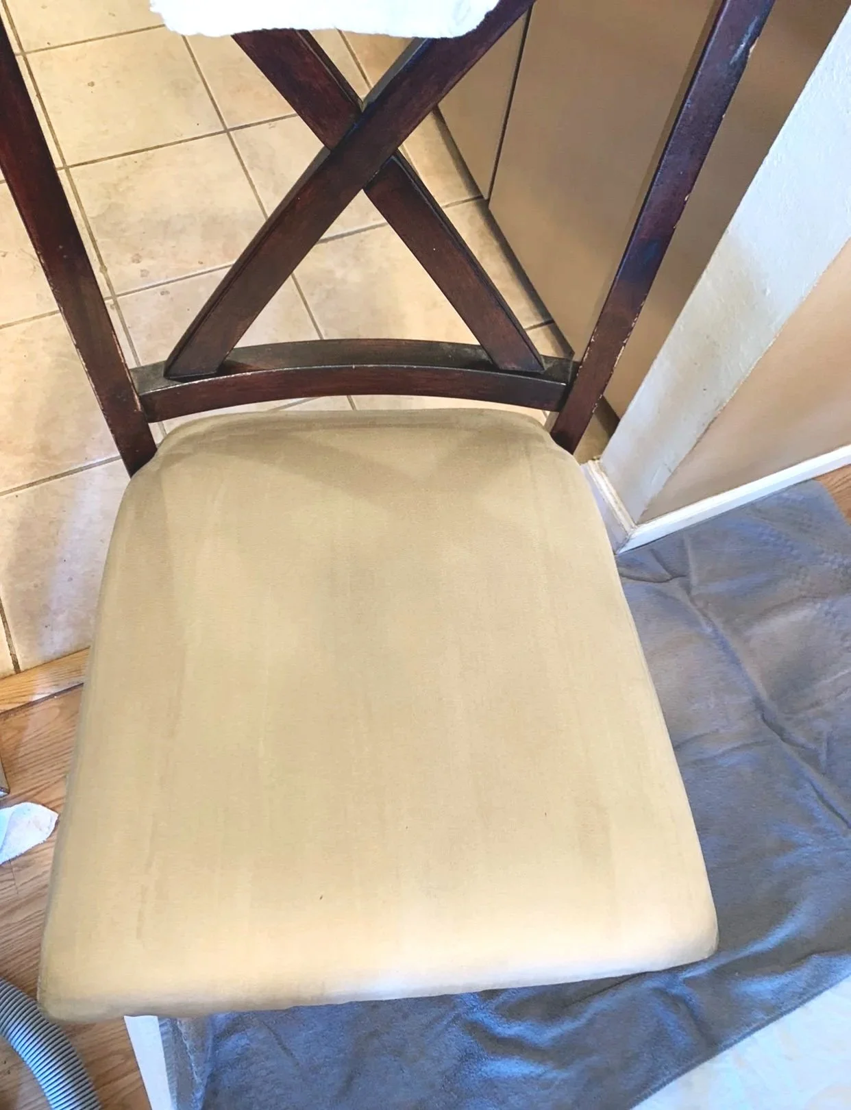 A wooden chair with a beige upholstered seat, positioned on a tiled kitchen floor next to a countertop and cabinets.