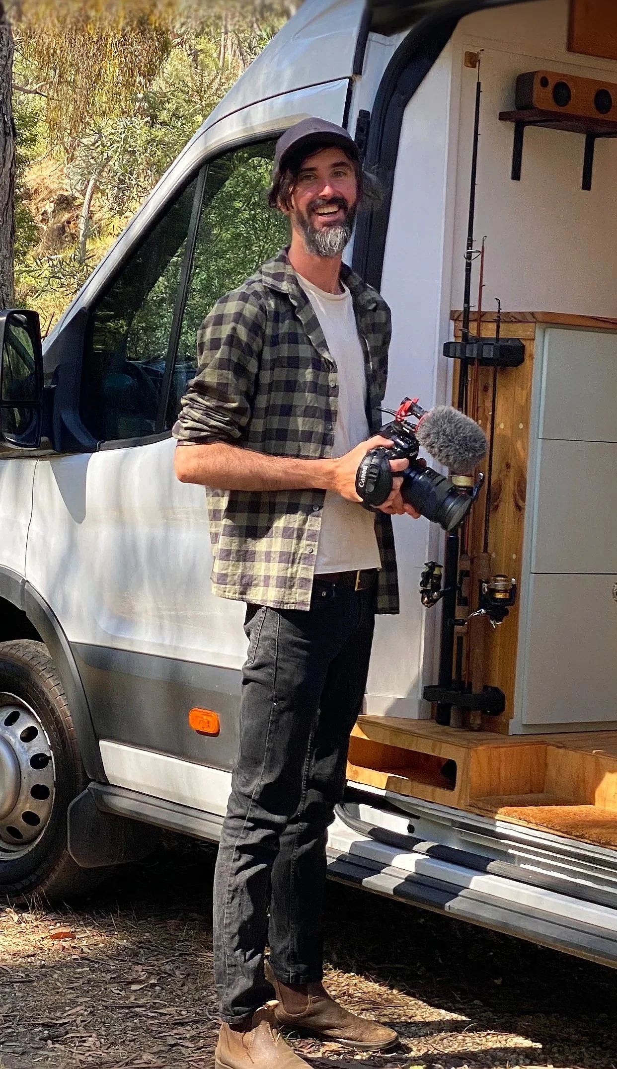 A man with a beard holding a camera with a microphone, standing outside a white van with a wooded background.