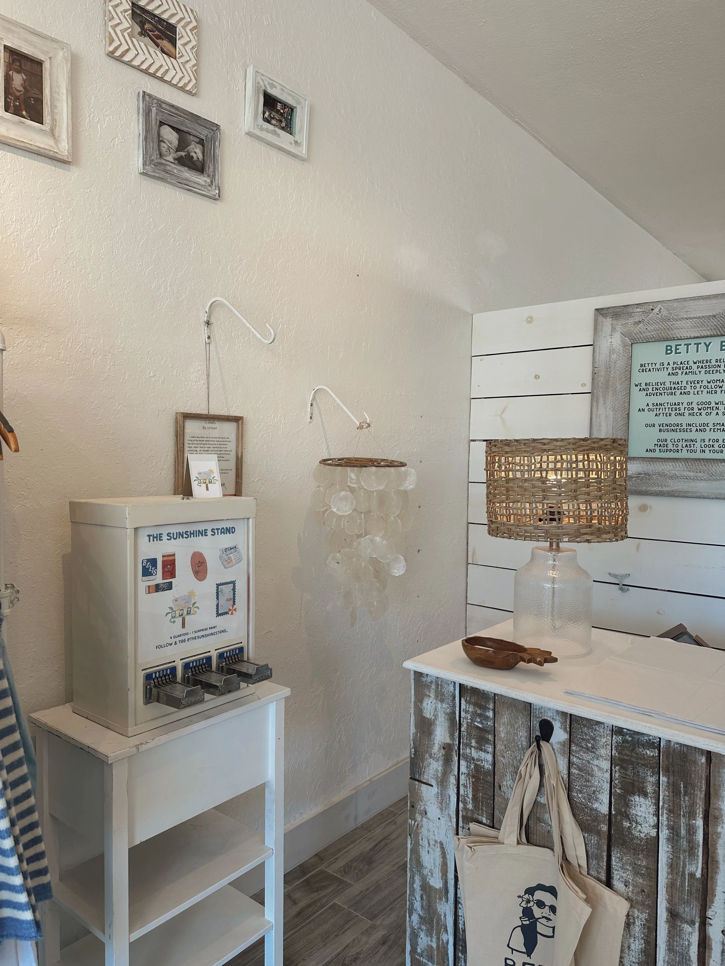 A corner of a room with a white wall decorated with framed photos, a Sunshine Stand beverage dispenser, a shell-shaped chandelier, a rustic wooden table with a glass vase lamp, a small wooden tray, and a tote bag hanging on the side of the table.