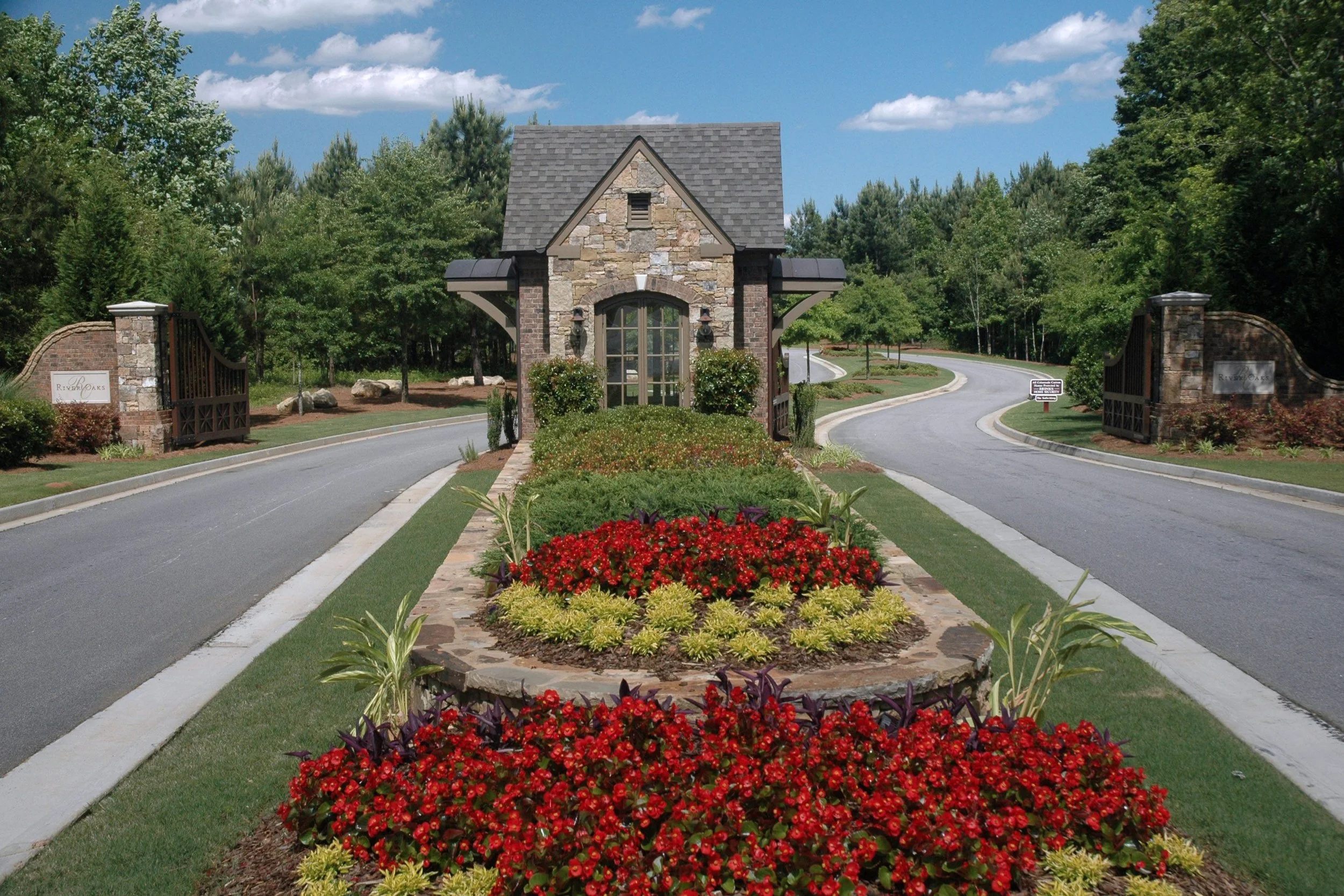 Entrance to a gated community with stone and brick gatehouse, colorful flowerbeds with red, yellow, and green plants in the center, and a curved road surrounded by trees.