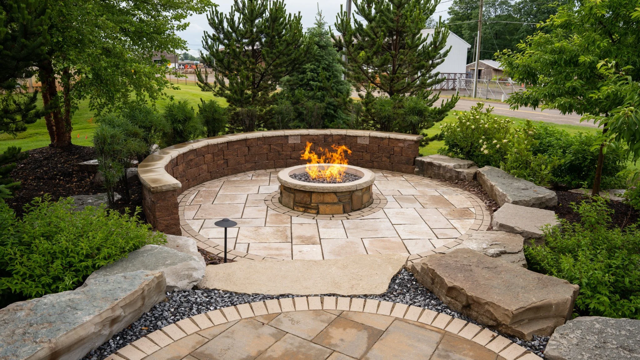 A landscaped backyard area with a circular stone fire pit surrounded by a stone patio, a curved brick retaining wall, and lush green trees and shrubs.