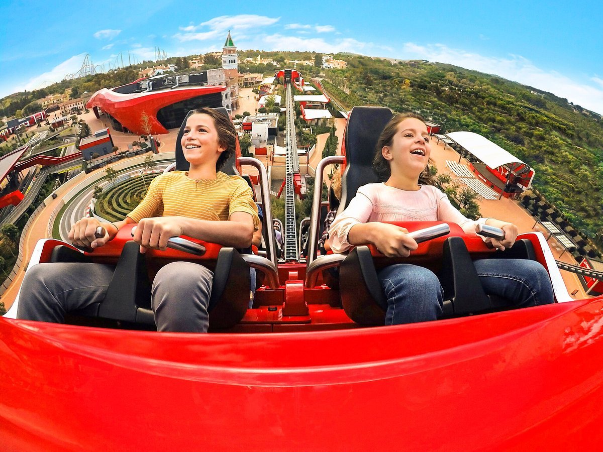 Two girls enjoying a roller coaster ride at an amusement park, with a scenic background of trees and buildings.
