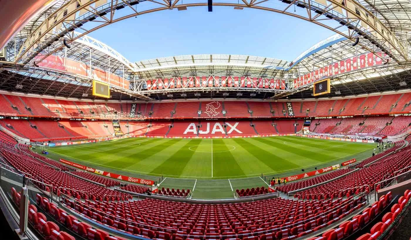 Wide-angle view of a nearly empty soccer stadium with red seats, a green field, and a bright blue sky overhead. The stadium has 'AJAX' displayed prominently on the seats and various advertisements along the sidelines.