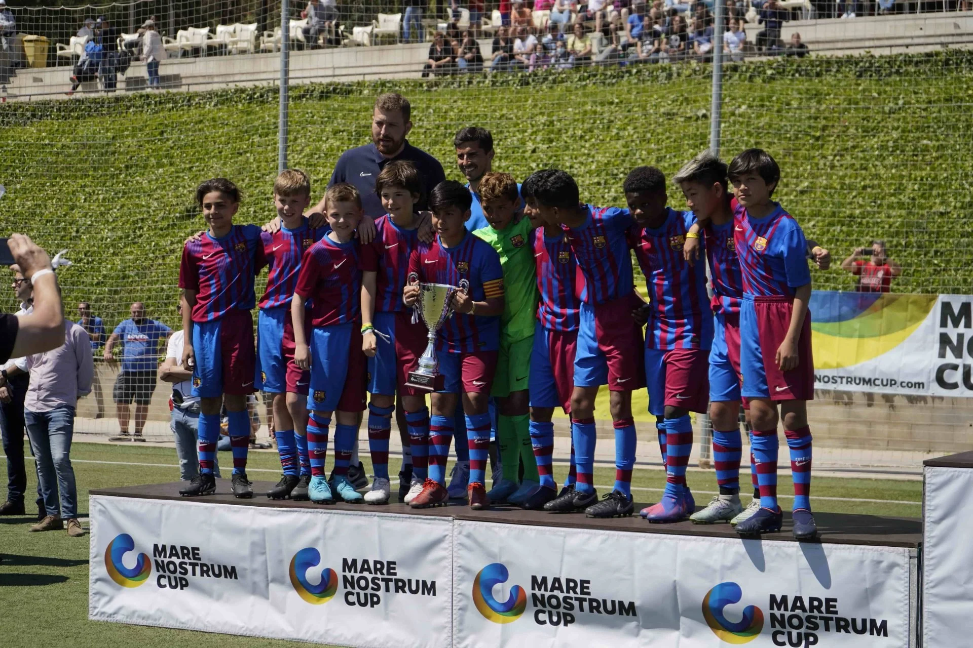 Children in soccer uniforms posing on a winners' podium with a trophy after a youth soccer tournament, with spectators in the background.