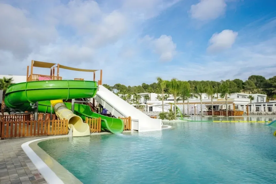 Water park with green and yellow slides, some enclosed and some open, next to a large swimming pool surrounded by palm trees and lounge chairs, under a partly cloudy sky.
